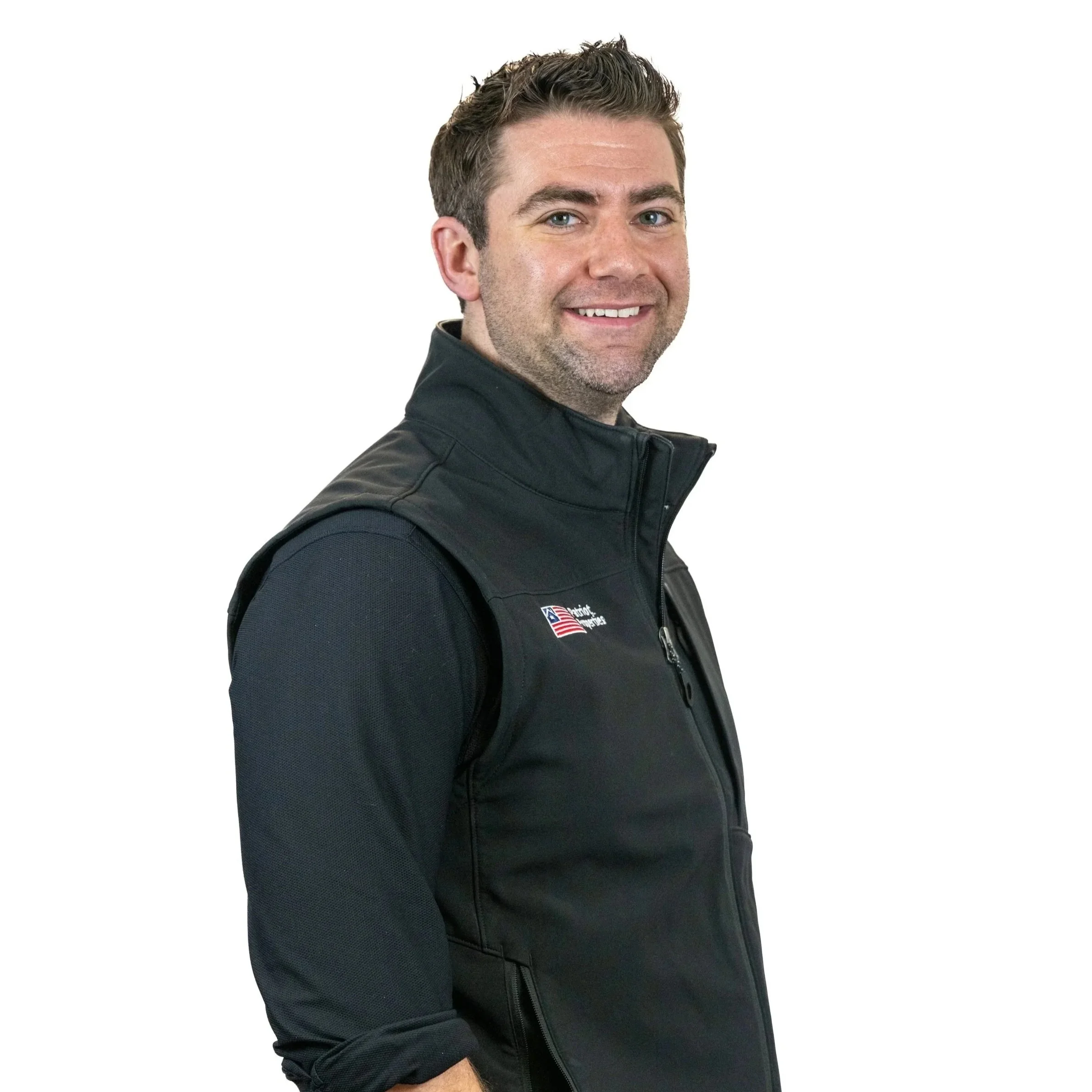 A smiling man with short brown hair and a beard, wearing a black vest with an American flag logo, standing against a white background.