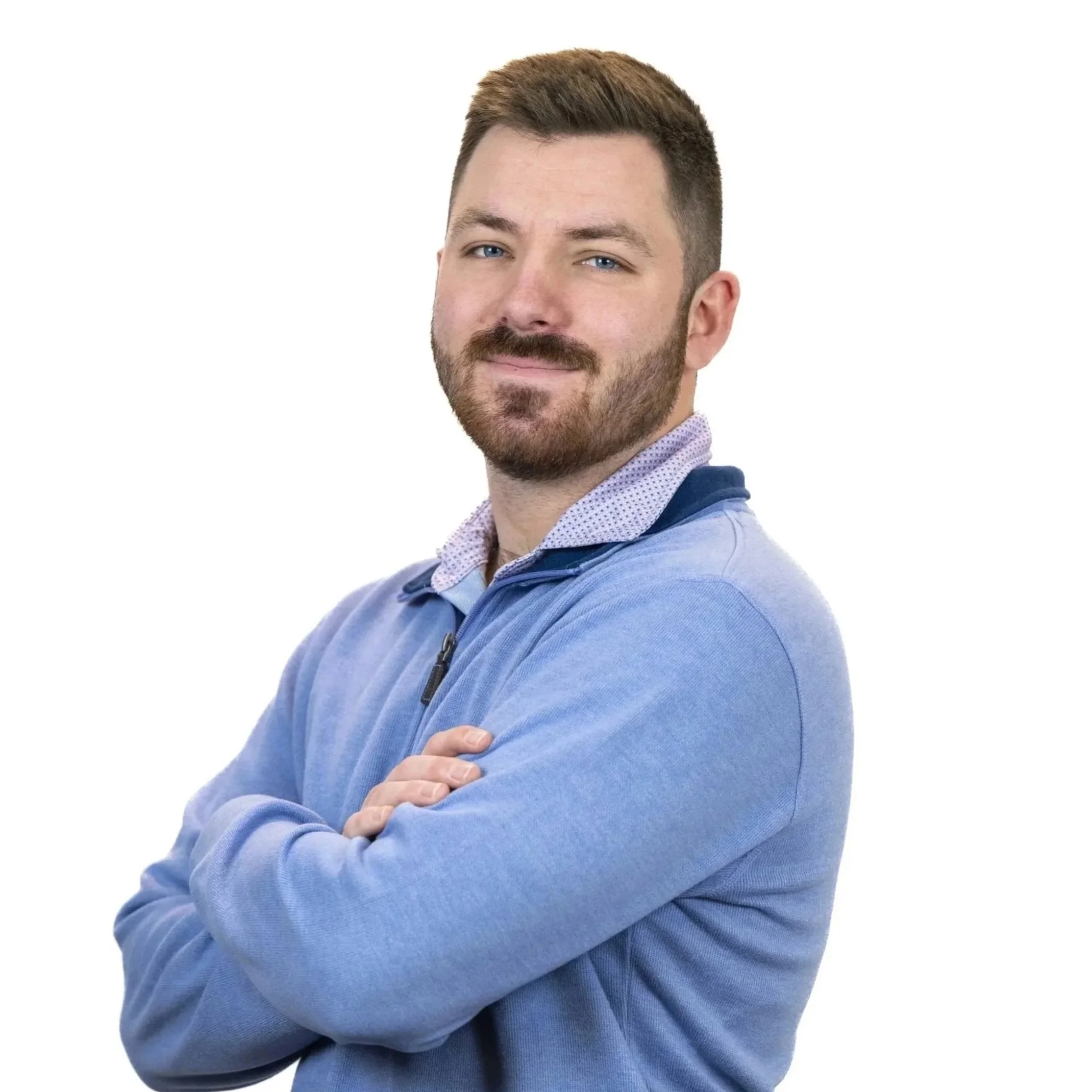A man with light skin, beard, and short brown hair, wearing a light blue zip-up sweater with a collared shirt underneath, standing with arms crossed and smiling slightly against a white background.