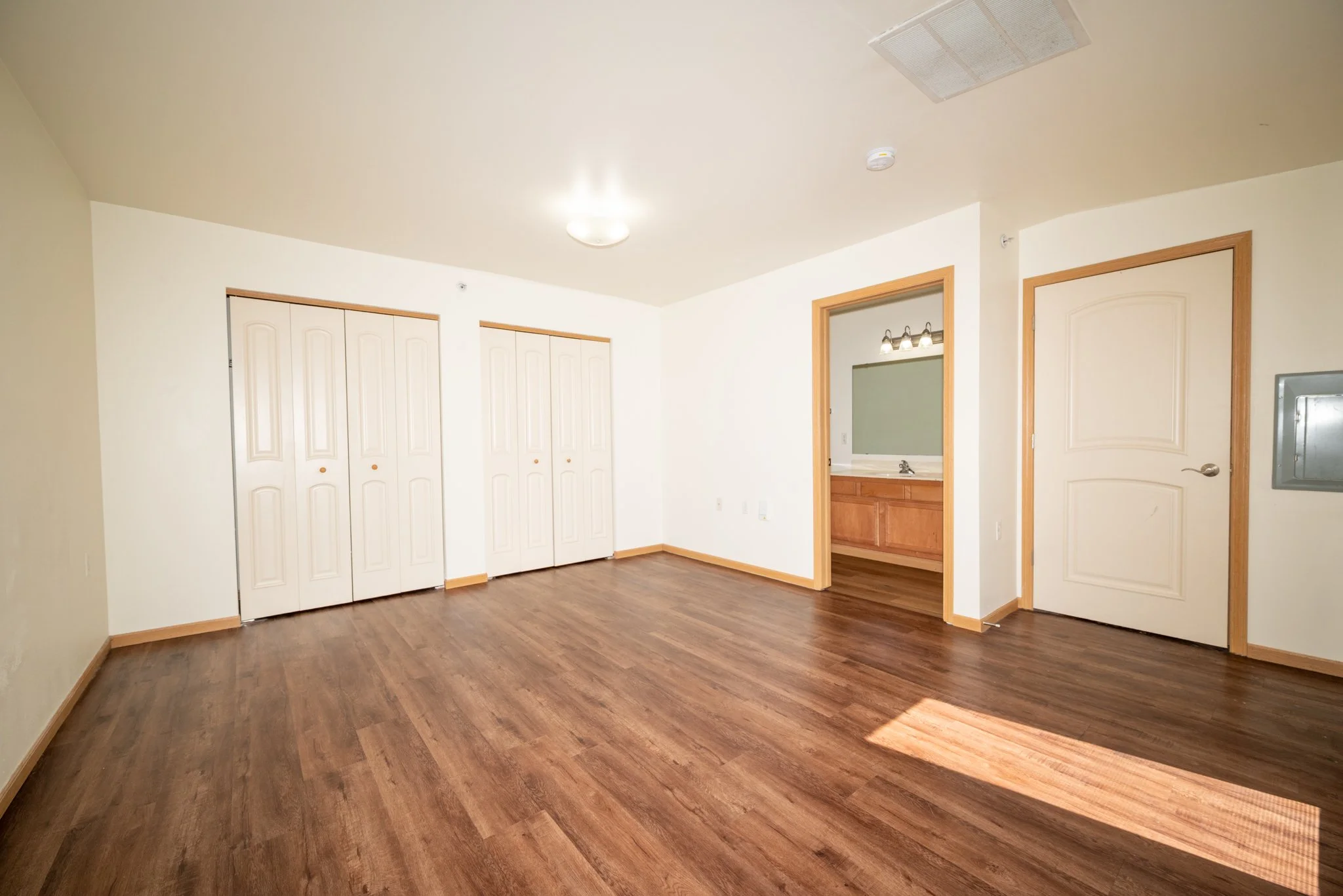 Empty room with hardwood floors, white walls, two closet doors, a small wall mirror, and a doorway leading to a bathroom or kitchen area with wooden cabinetry and a mirror with lights above it.