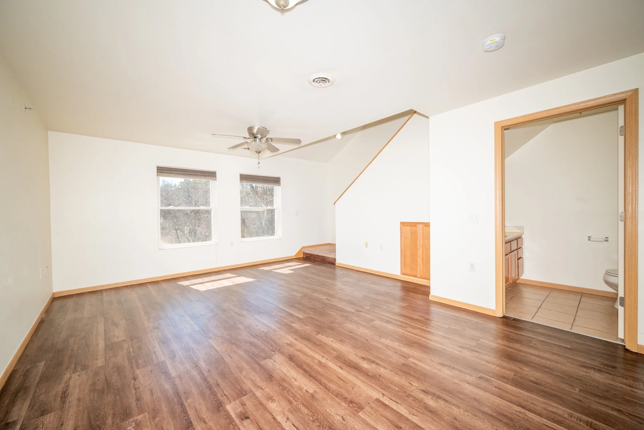 Empty living area with wood floors, two large windows, ceiling fan, and room divider leading to a bathroom with wooden vanity.