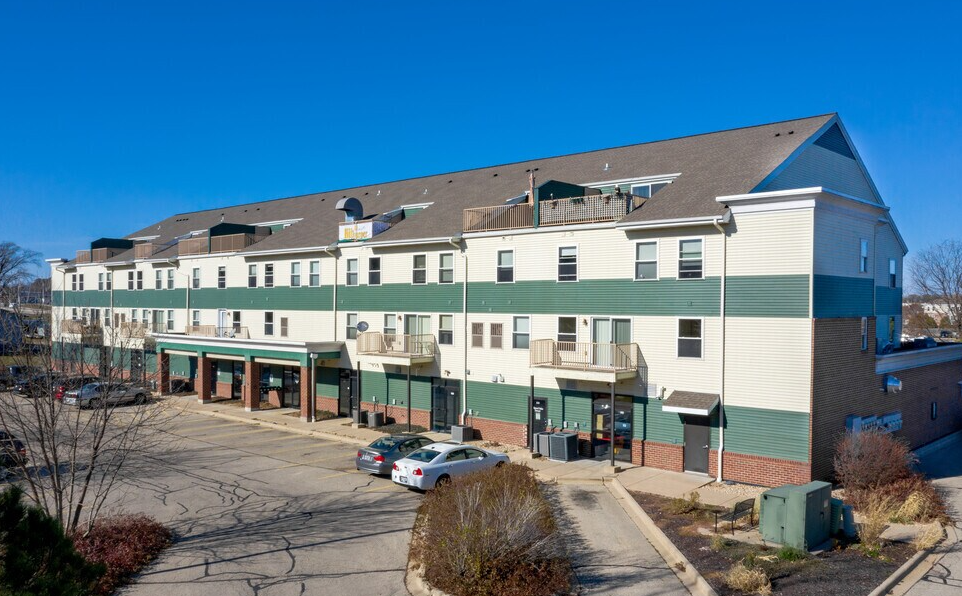 A multi-story apartment building with green and white exterior, balconies, and a parking lot with parked cars in front.
