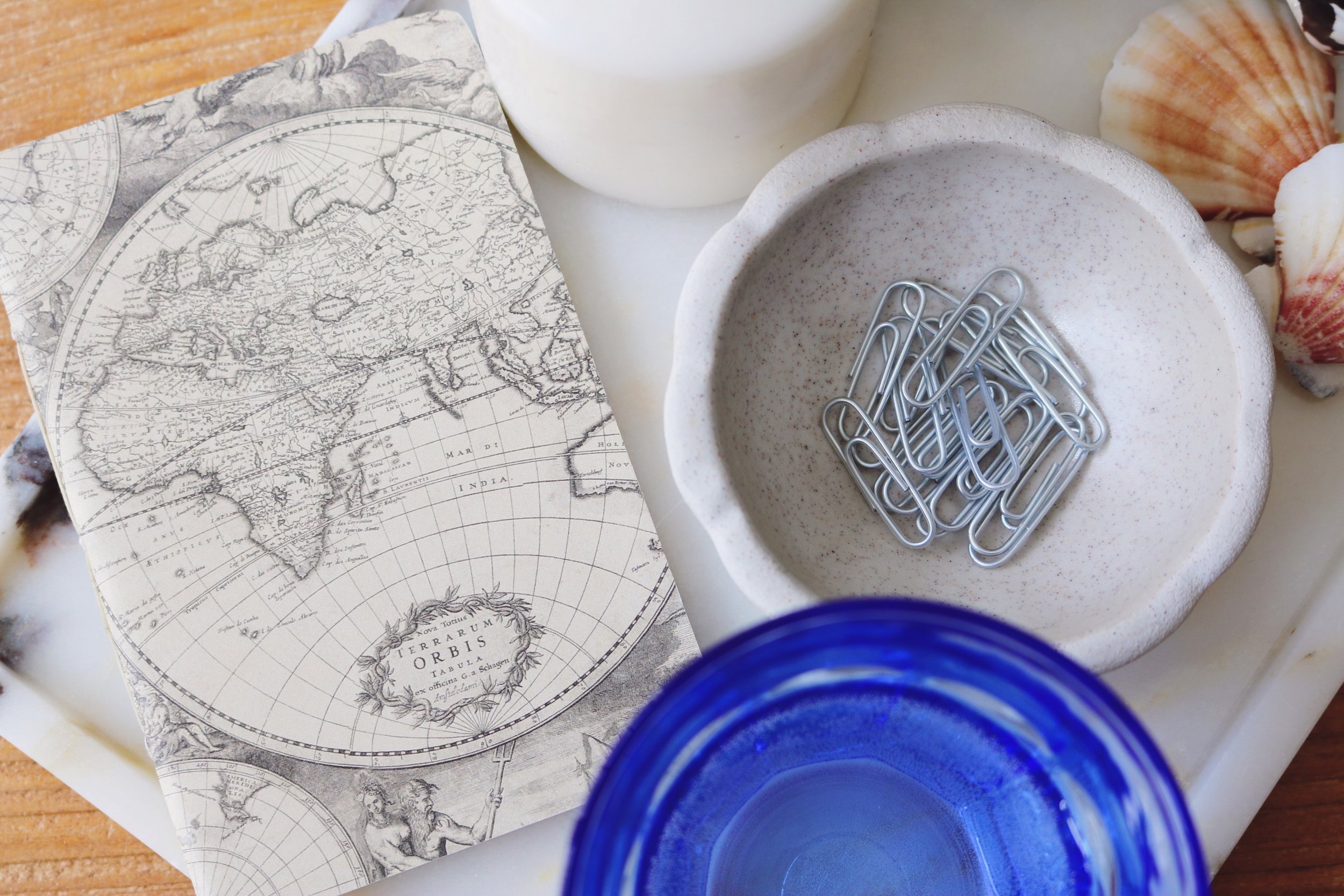 A mini notebook with an old map of the world printed on the front is laid atop a while marble tray with splashes of browns and grays. Next to it a small ceramic dish with the edge of a paper clip and a cobalt blue water glass in the lower corner.