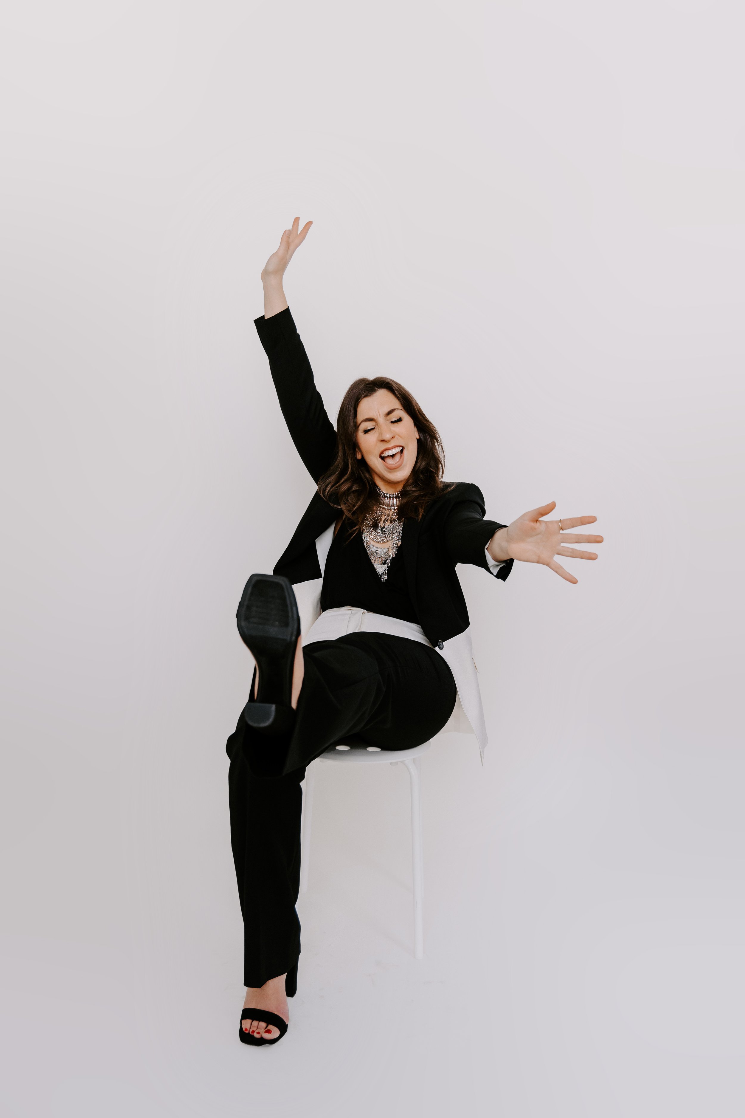 Our founder dressed in a black suit and heels. They are seated on a stool mid-laugh with their arms outstretched and hands open. One of their legs is kicked into the air showing off their personality and humor.
