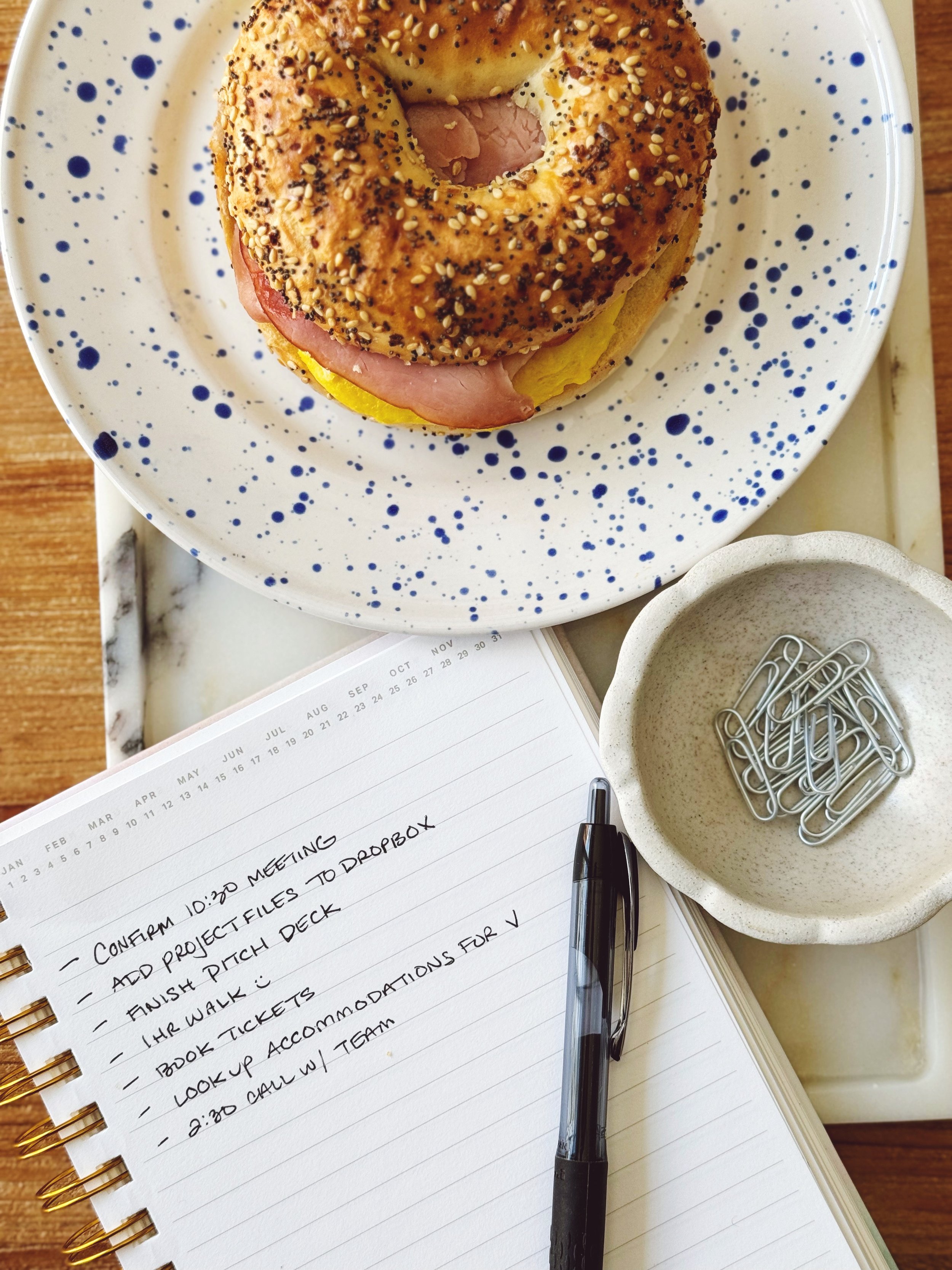 A white plate with blue speckles holds a breakfast bagel. It sits atop a marble tray that also has a notebook with a list of to-do's and a small ceramic dish with paperclips.