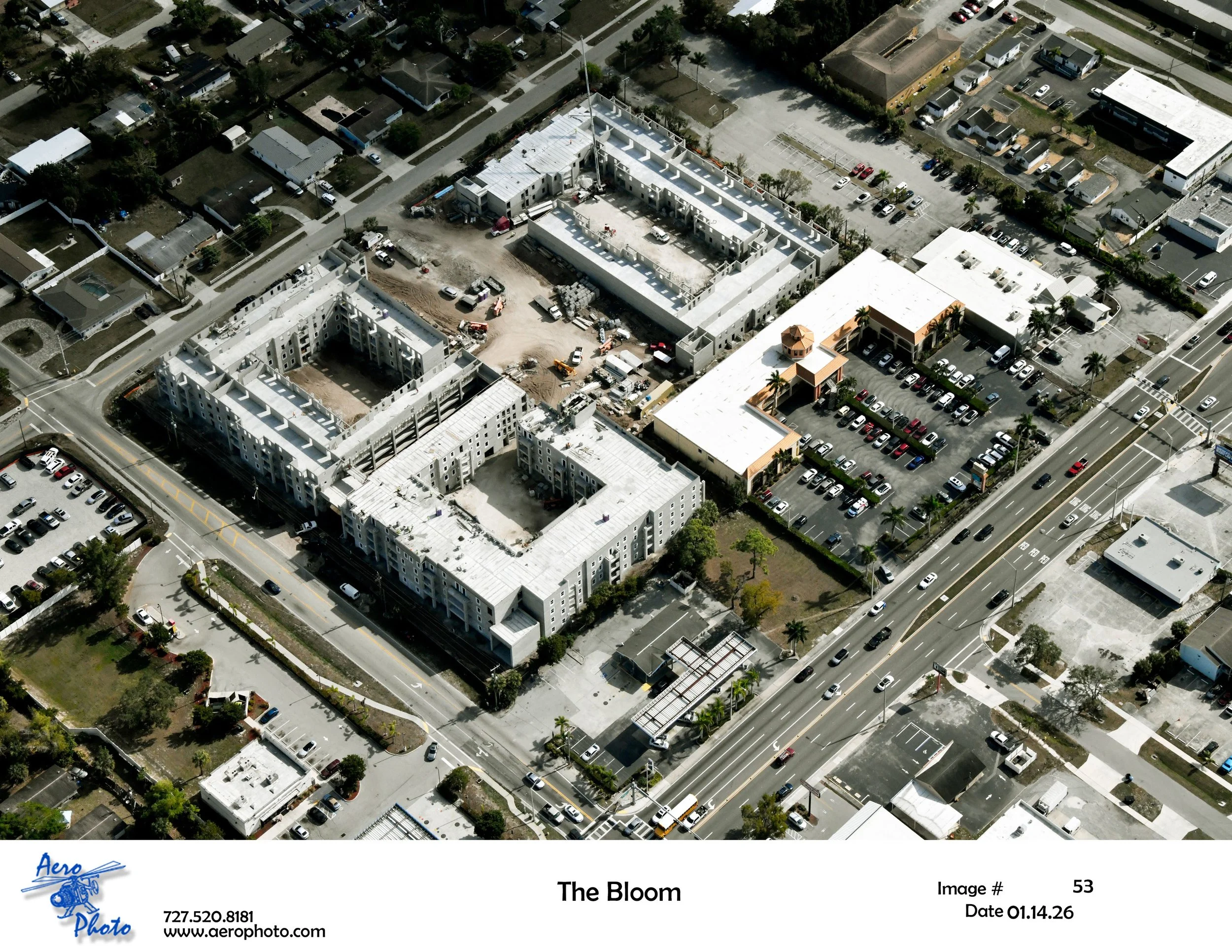 Aerial view of a construction site with partially built multi-story buildings, surrounding parking lots, and nearby roads with cars. Residential neighborhood and commercial buildings are visible in the background.