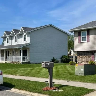 Suburban neighborhood with two houses, manicured lawns, a gray mailbox in the foreground, and clear blue sky.