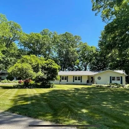 Single-story yellow house with a spacious front lawn, surrounded by trees and shrubs under a clear blue sky.