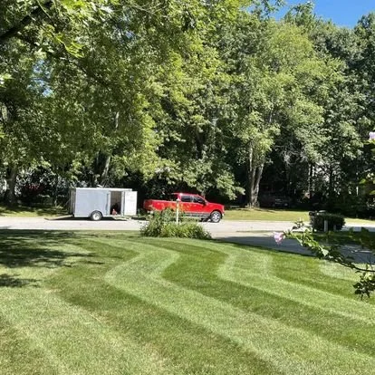 Manicured lawn with striped mowing pattern, a red truck, and white trailer on a suburban street with trees in the background.
