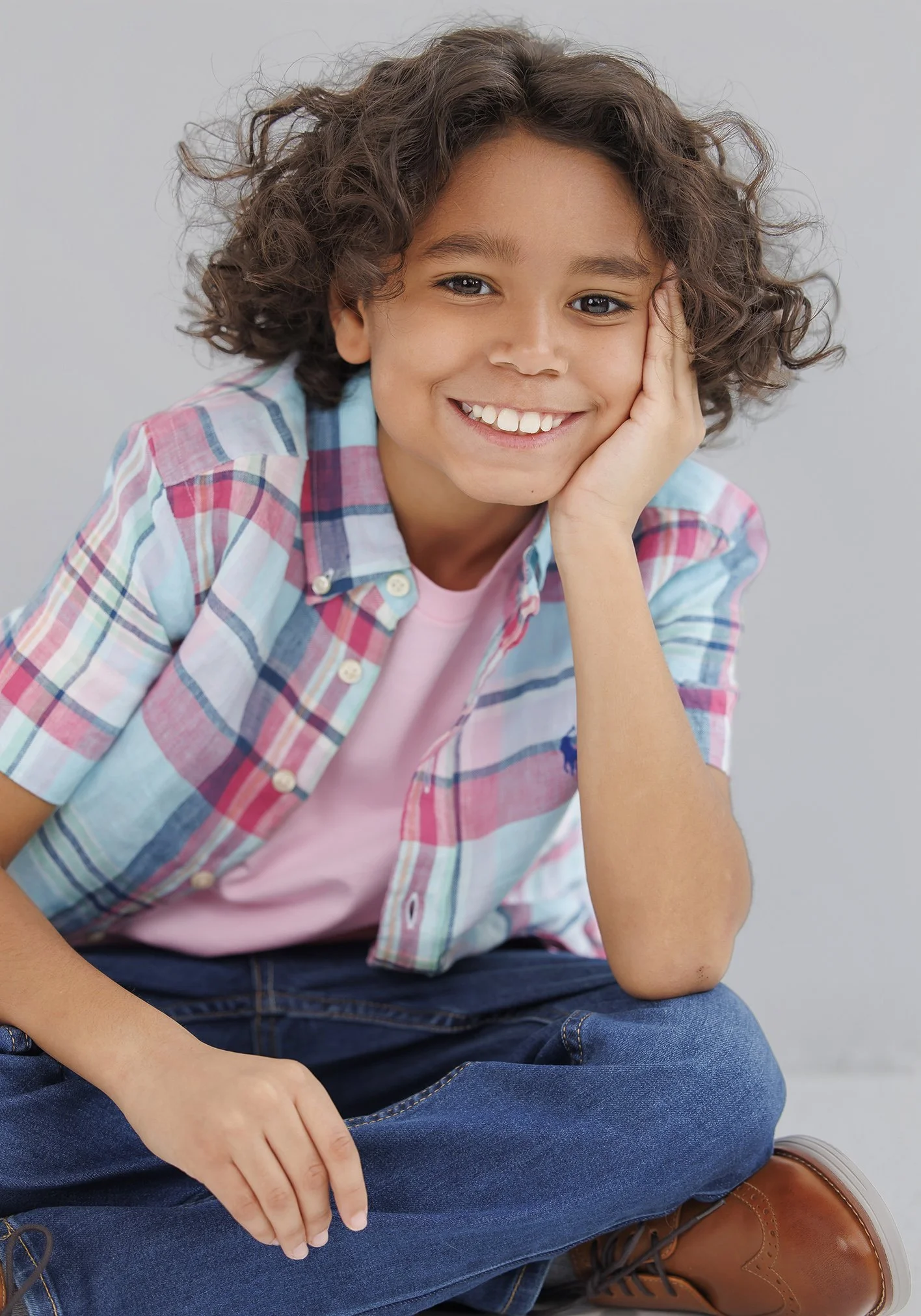 A young boy with curly hair smiling, sitting cross-legged with one hand resting on his face, wearing a pink t-shirt, plaid shirt, jeans, and brown shoes.