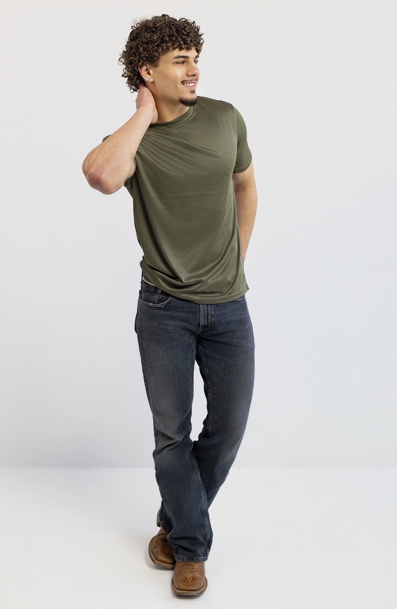Young man wearing a green t-shirt, dark jeans, and brown shoes, standing with one hand behind his head, smiling, against a plain white background.