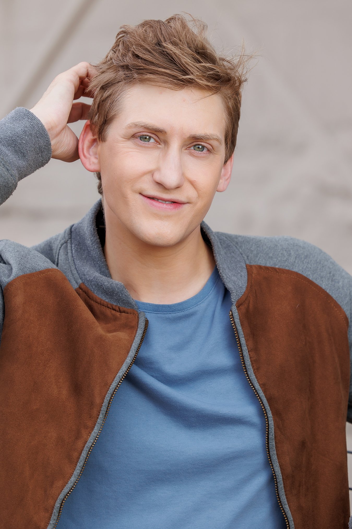 A young man with light brown hair styled with a quiff, wearing a blue t-shirt and a brown and gray jacket, standing outdoors against a blurred gray background. He is smiling with a slight smirk and has his right hand touching his hair.