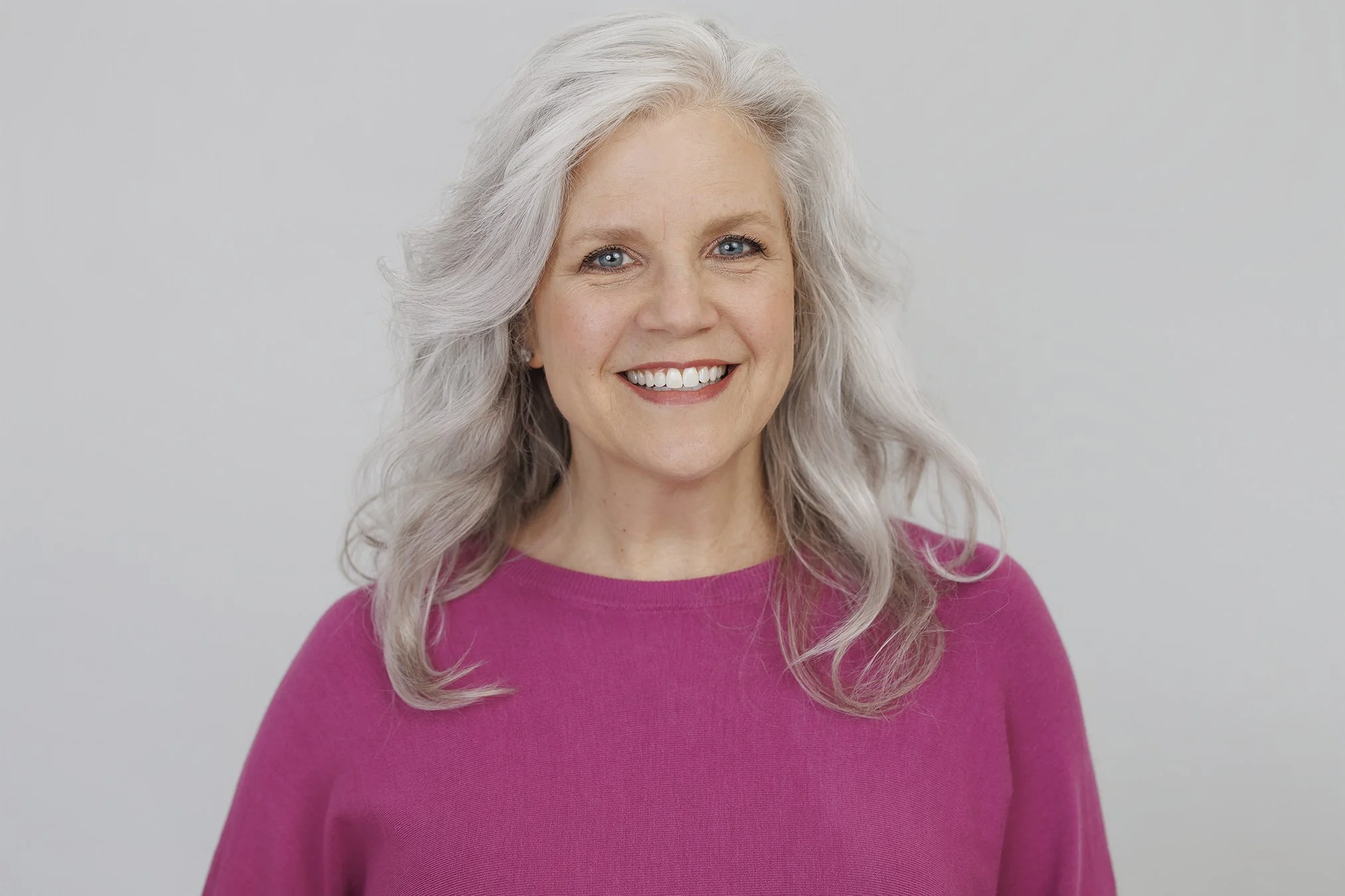 Portrait of an older woman with long, wavy gray hair, smiling and wearing a bright pink top, standing against a plain white background.
