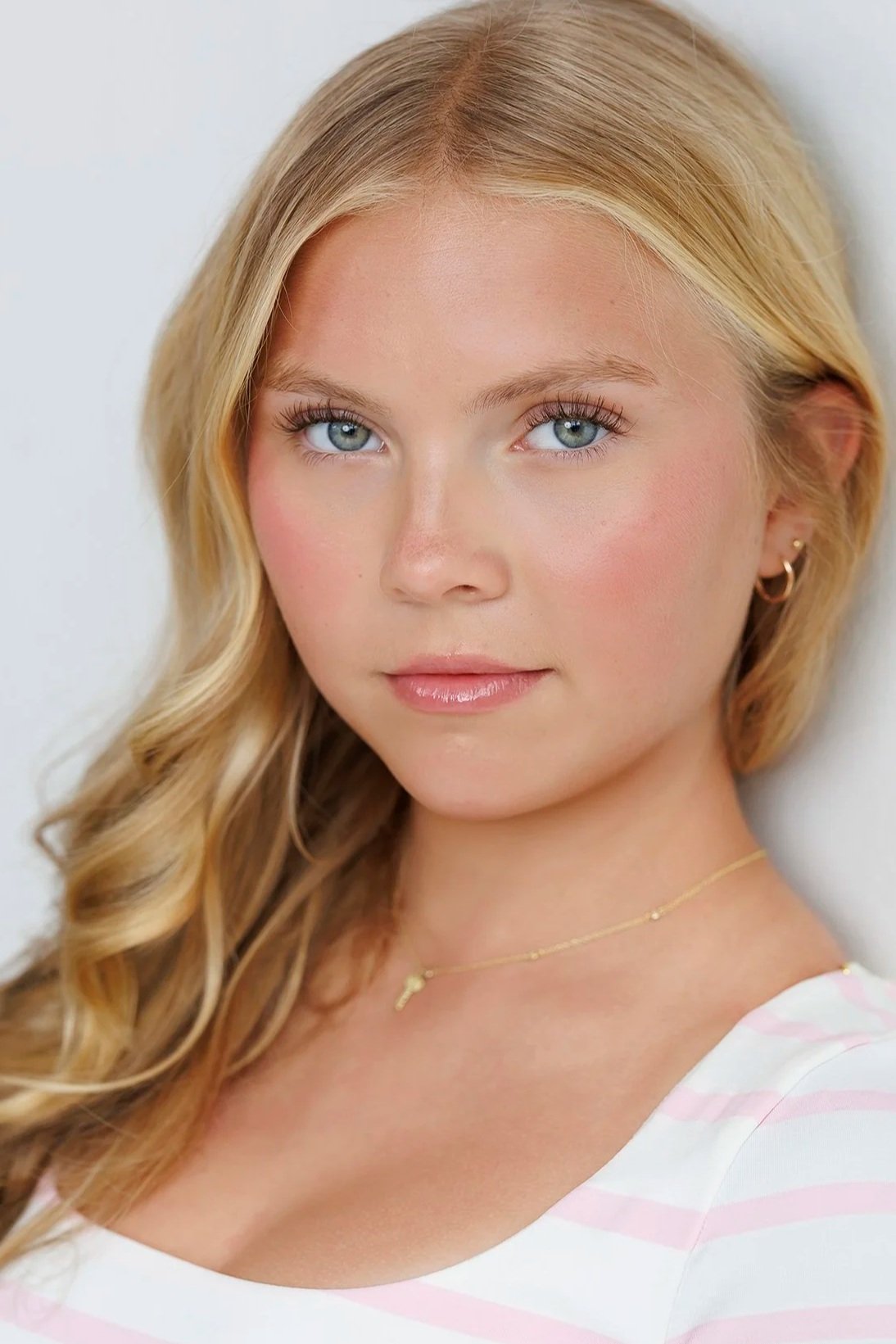 Close-up portrait of a young woman with blonde hair, blue eyes, and light makeup, wearing a gold necklace and earrings, with a white striped top, standing against a plain white background.