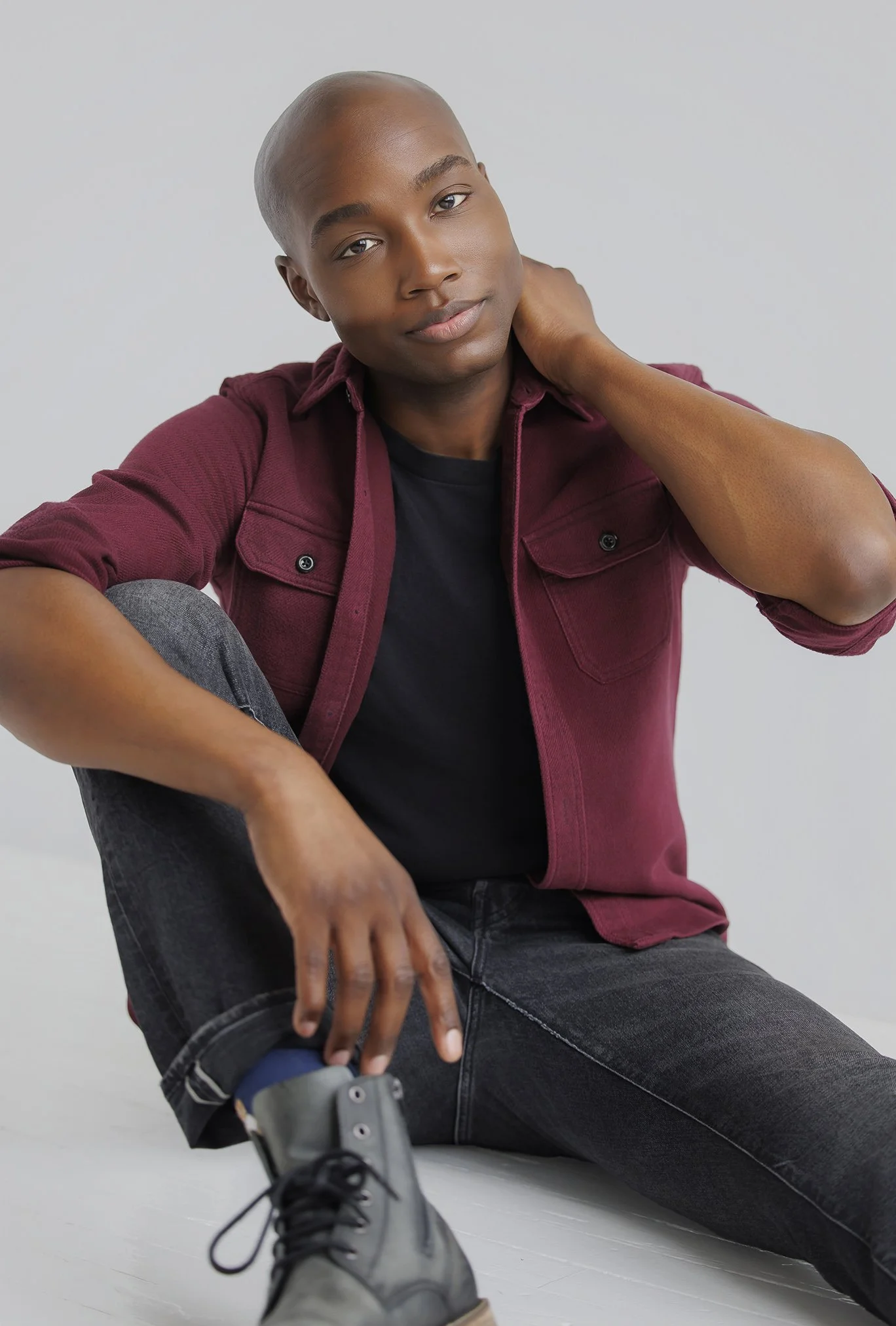 Young man sitting on floor in casual clothing, wearing a maroon shirt and black jeans, with one hand on his ankle and the other touching his neck, against a plain light background.