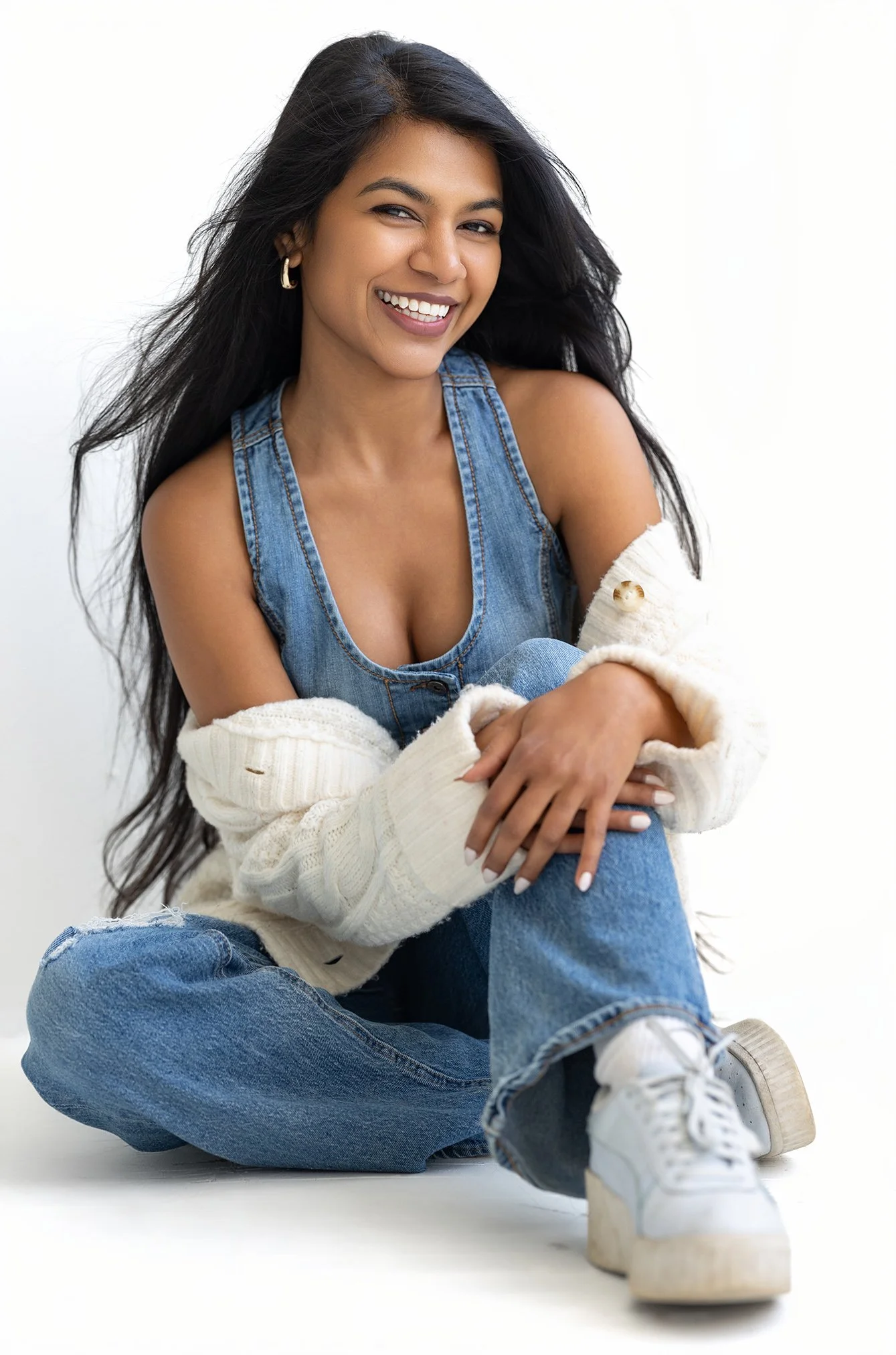 A young woman with long dark hair, smiling, wearing a denim tank top, a cream knit sweater, ripped jeans, and platform sneakers, sitting cross-legged against a white background.