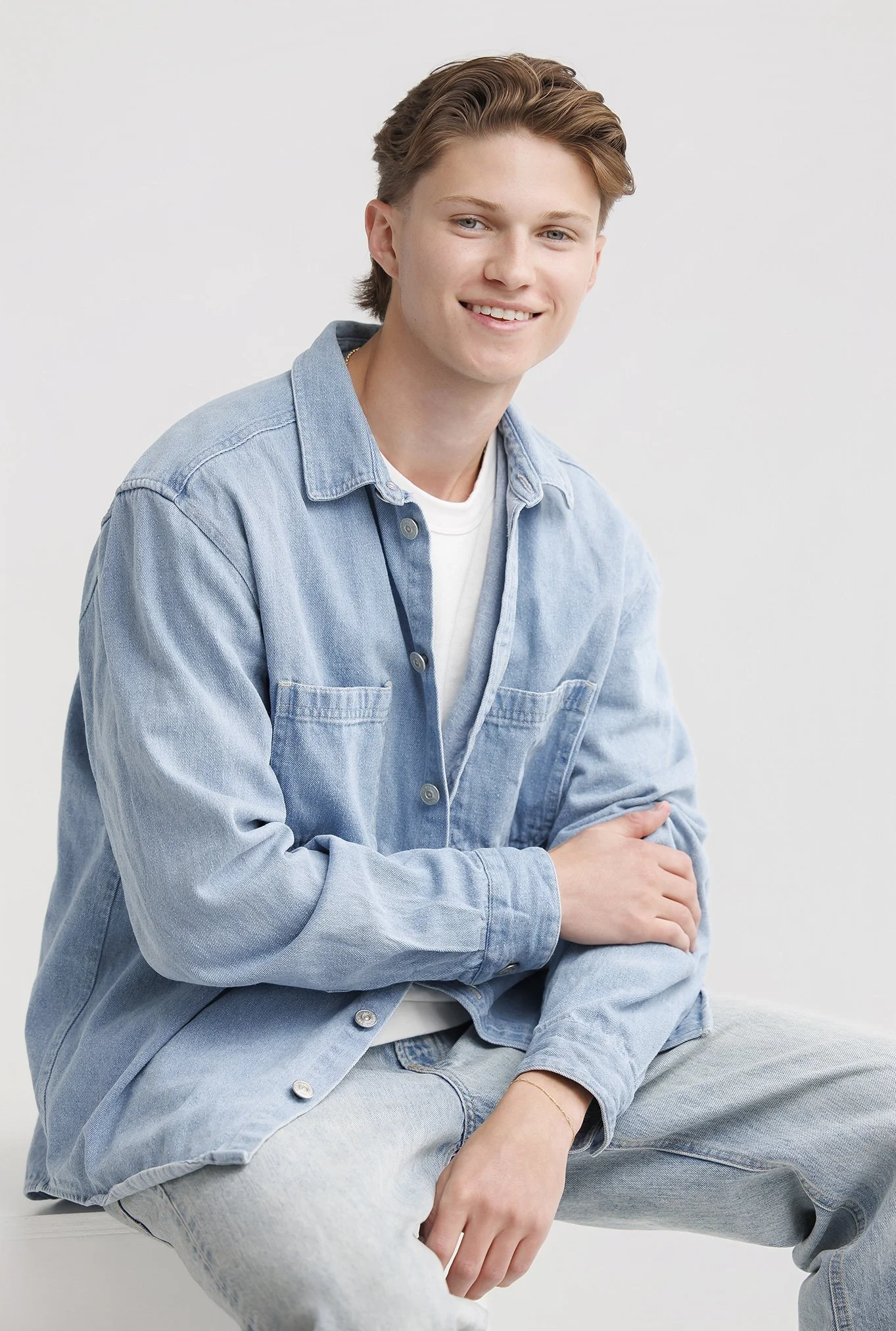 A young man with light brown hair, wearing a light blue denim jacket over a white t-shirt, smiling and sitting against a plain white background.