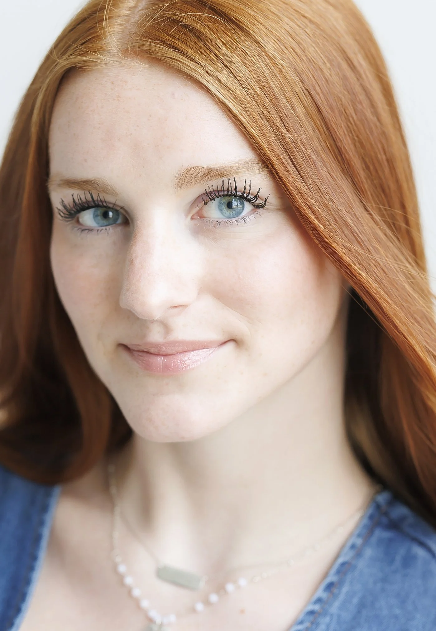 Close-up portrait of a young woman with red hair and blue eyes, wearing a denim top and layered necklaces, smiling softly.