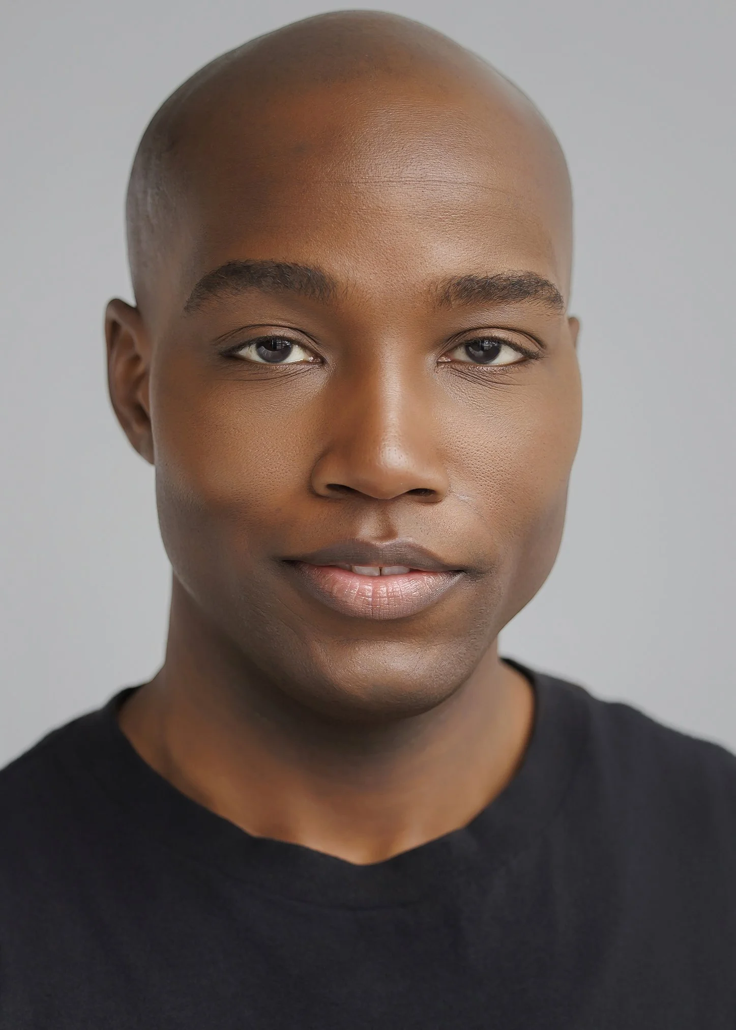Close-up portrait of a young African American man with a shaved head, neutral expression, wearing a black shirt, against a plain light gray background.