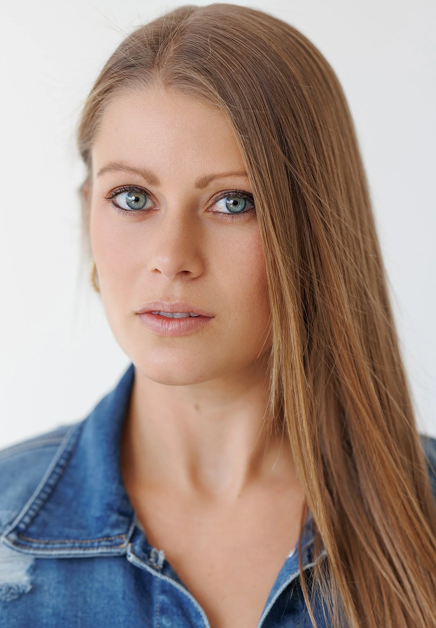 Close-up of a woman with long reddish-brown hair, blue eyes, and fair skin, wearing a denim jacket against a plain white background.