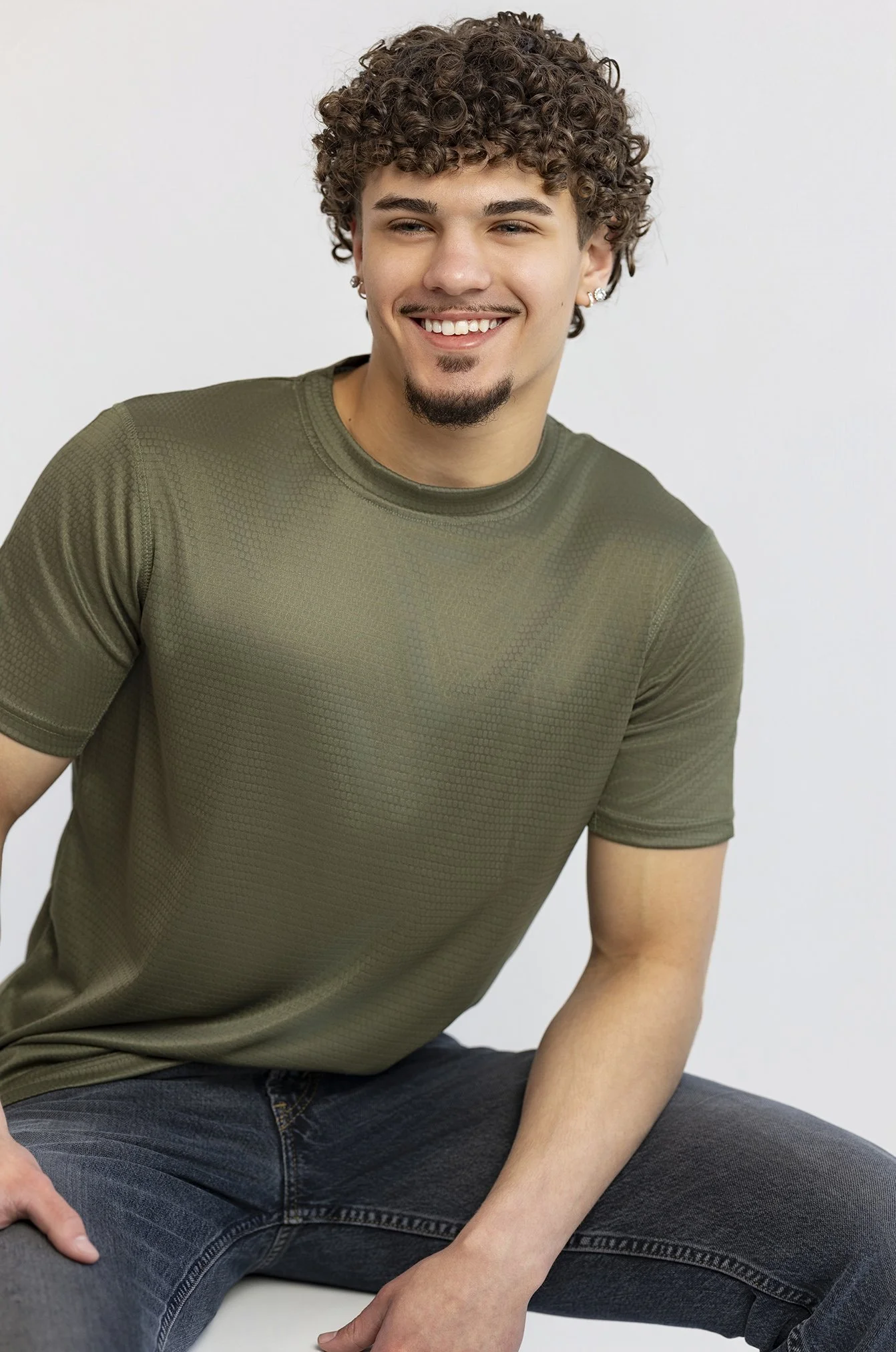 A young man with curly brown hair and a goatee, smiling and wearing a green textured t-shirt and dark jeans.