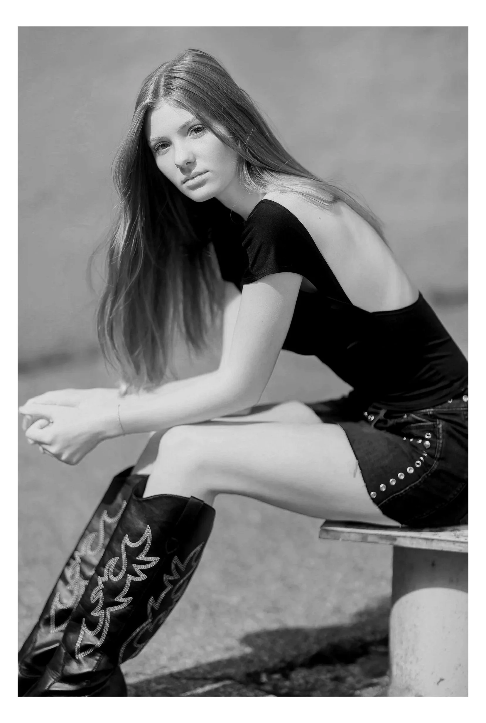 Black and white photo of a young woman sitting on a bench outdoors, wearing a black top with open shoulders, decorated shorts, and tall patterned boots, looking at the camera with a serious expression.