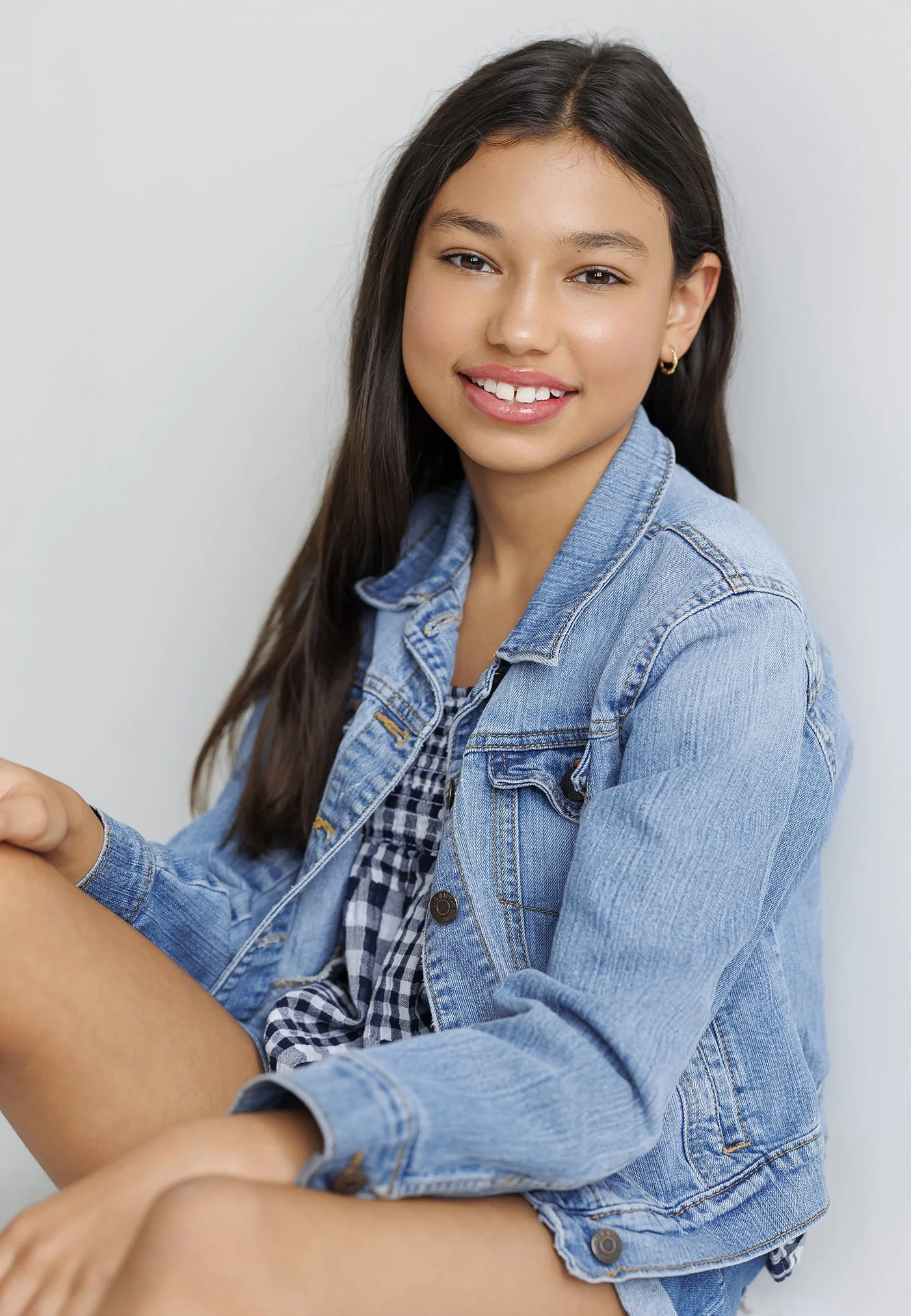 A young girl with long dark hair, wearing a denim jacket and a black-and-white checkered dress, smiling with white teeth in front of a plain light-colored background.
