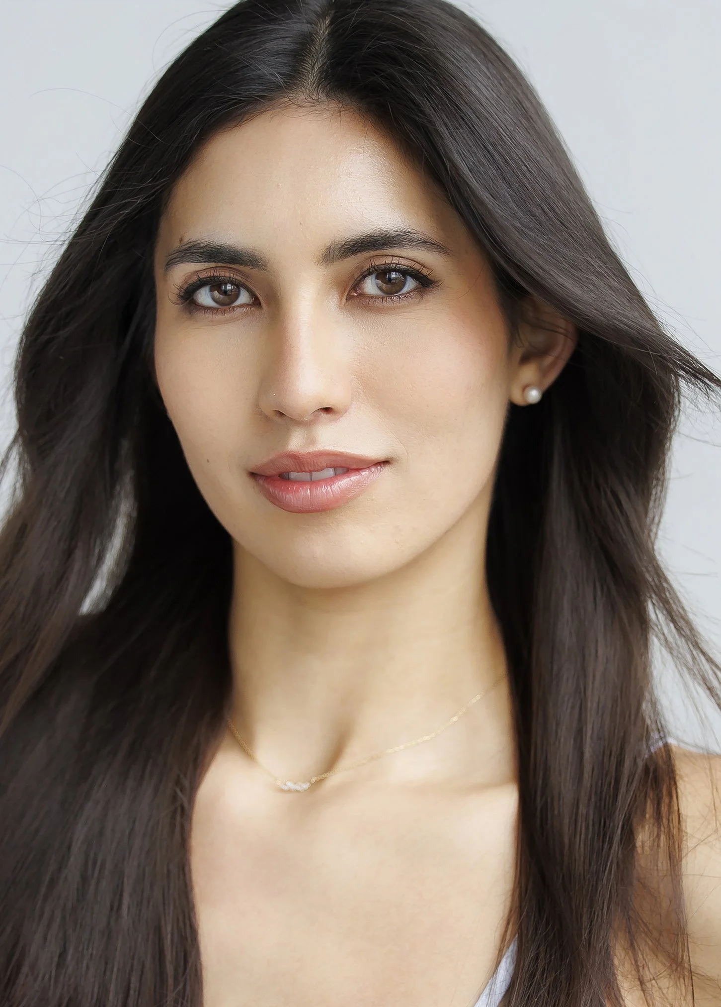 Close-up headshot of a woman with long brown hair, brown eyes, and fair skin, wearing pearl earrings and a delicate gold necklace, smiling softly.