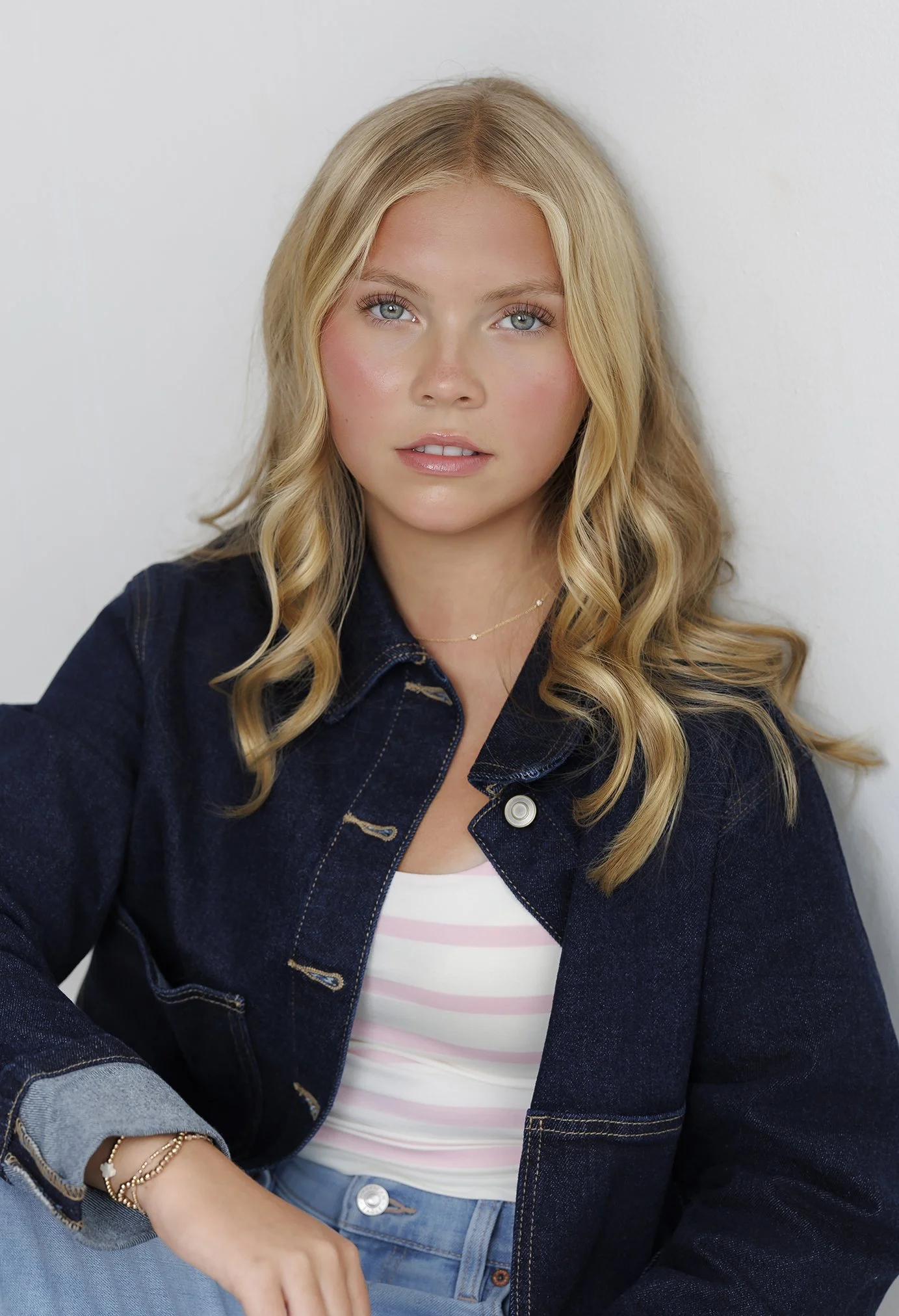 A young woman with blonde curly hair, wearing a denim jacket over a white and pink striped shirt, sitting against a white wall.