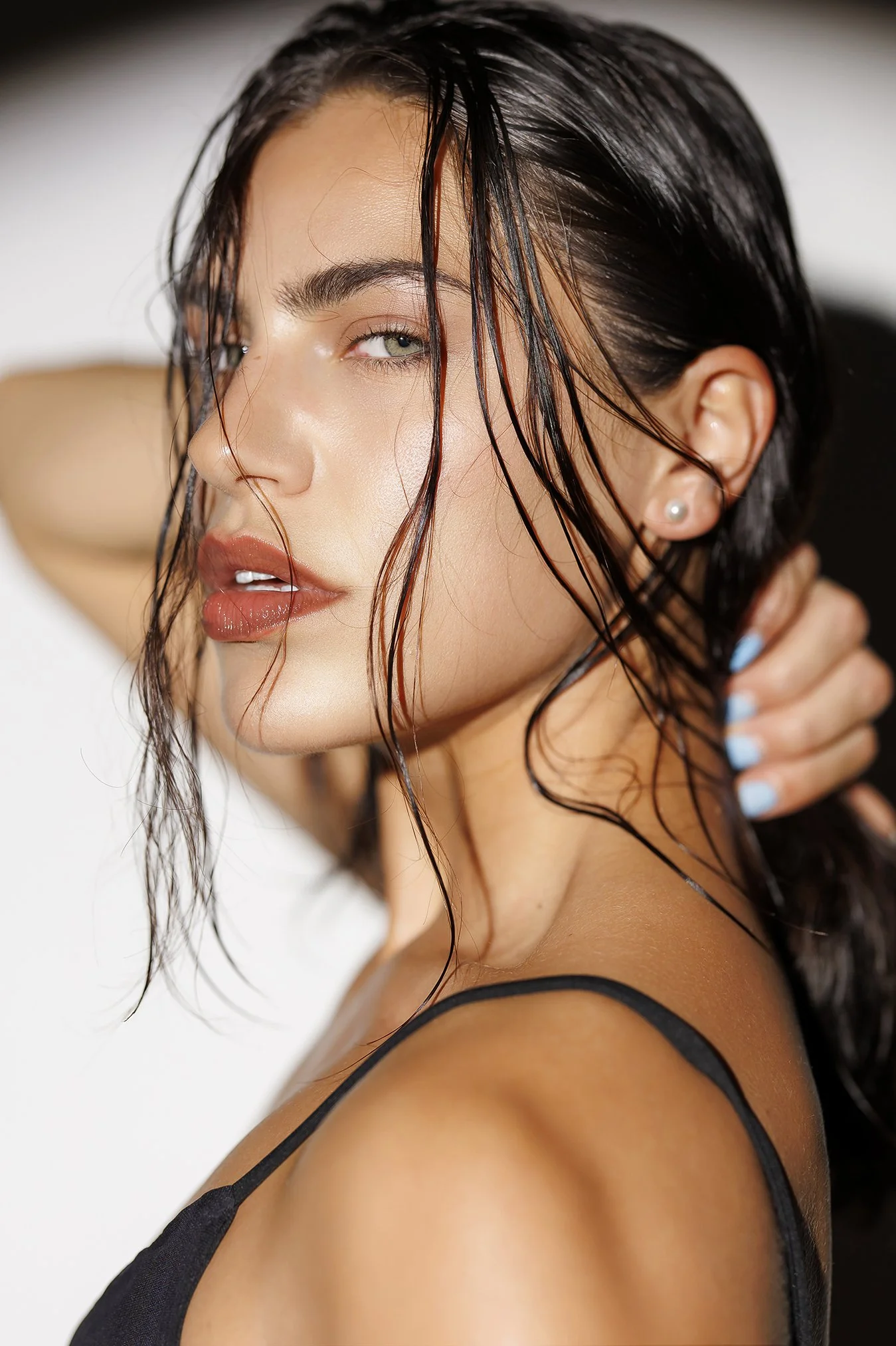 A young woman with wet, dark hair partially covering her face, wearing a black spaghetti strap top and pearl earrings, posing with her hand behind her neck and slightly parted lips.