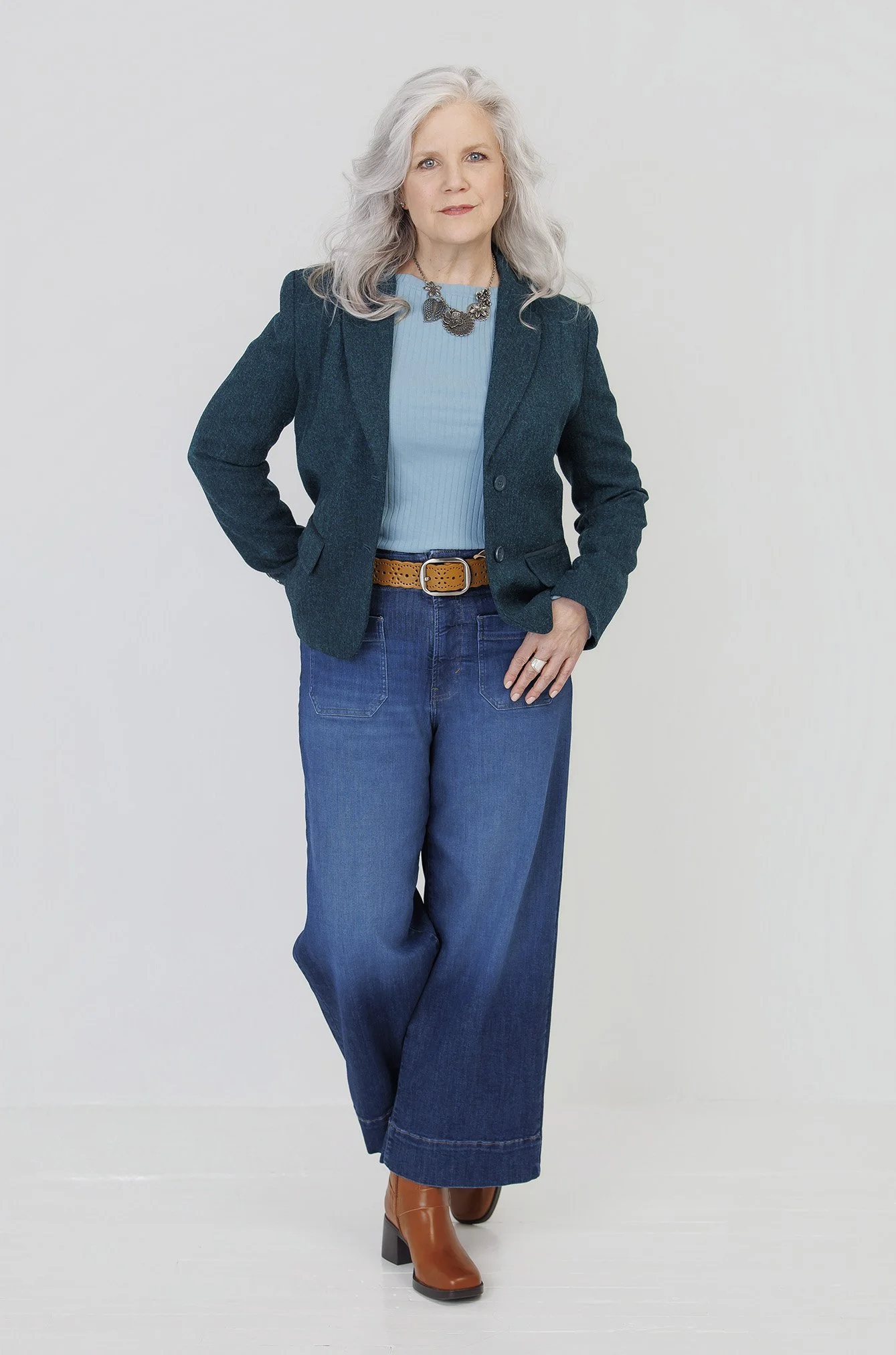 A woman with gray hair wearing a navy blazer, light blue top, wide-leg jeans, brown belt, and brown boots, standing against a plain white background.