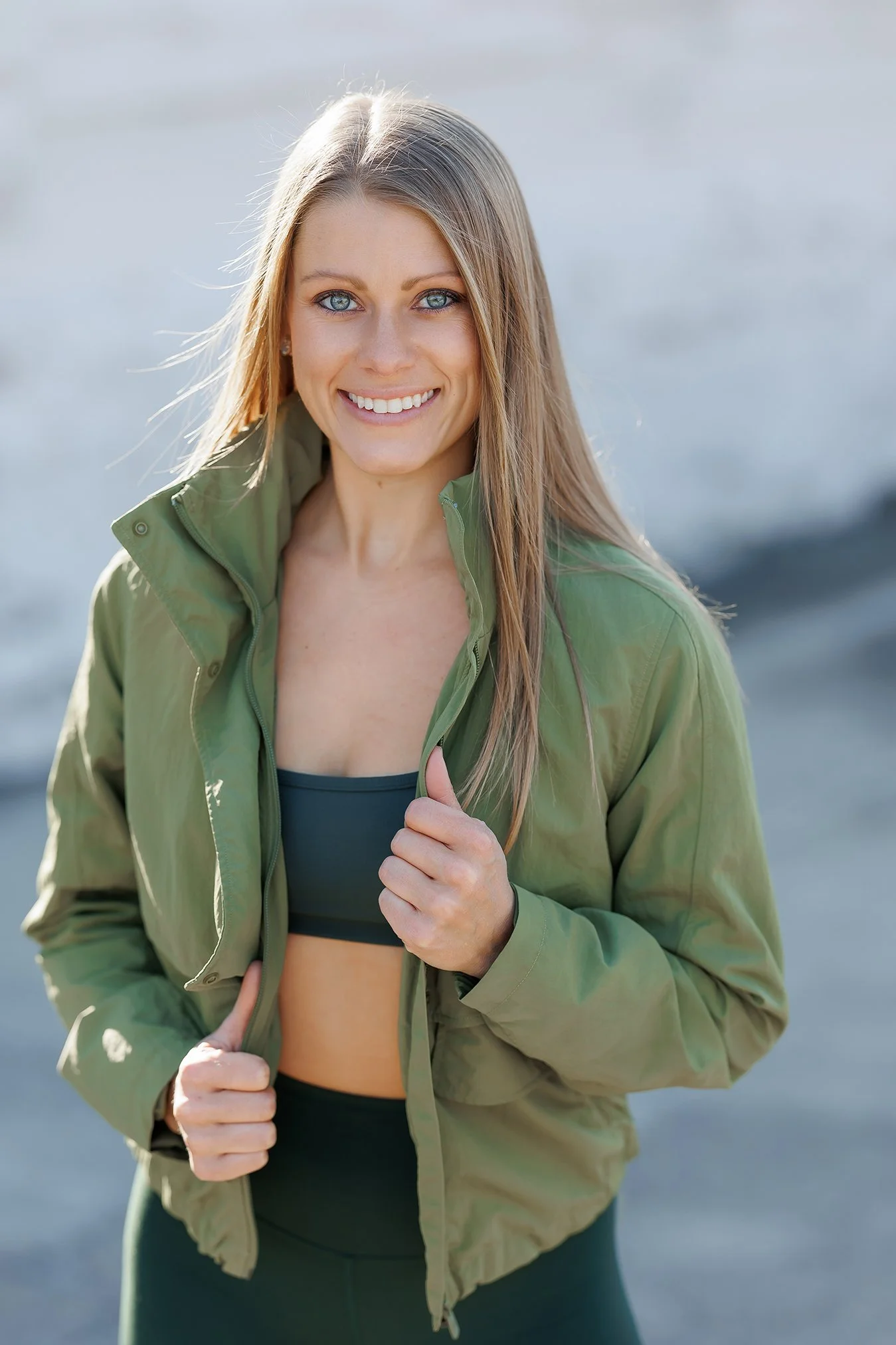 Woman smiling in athletic gear outdoors, wearing a green jacket and black sports bra, near water.