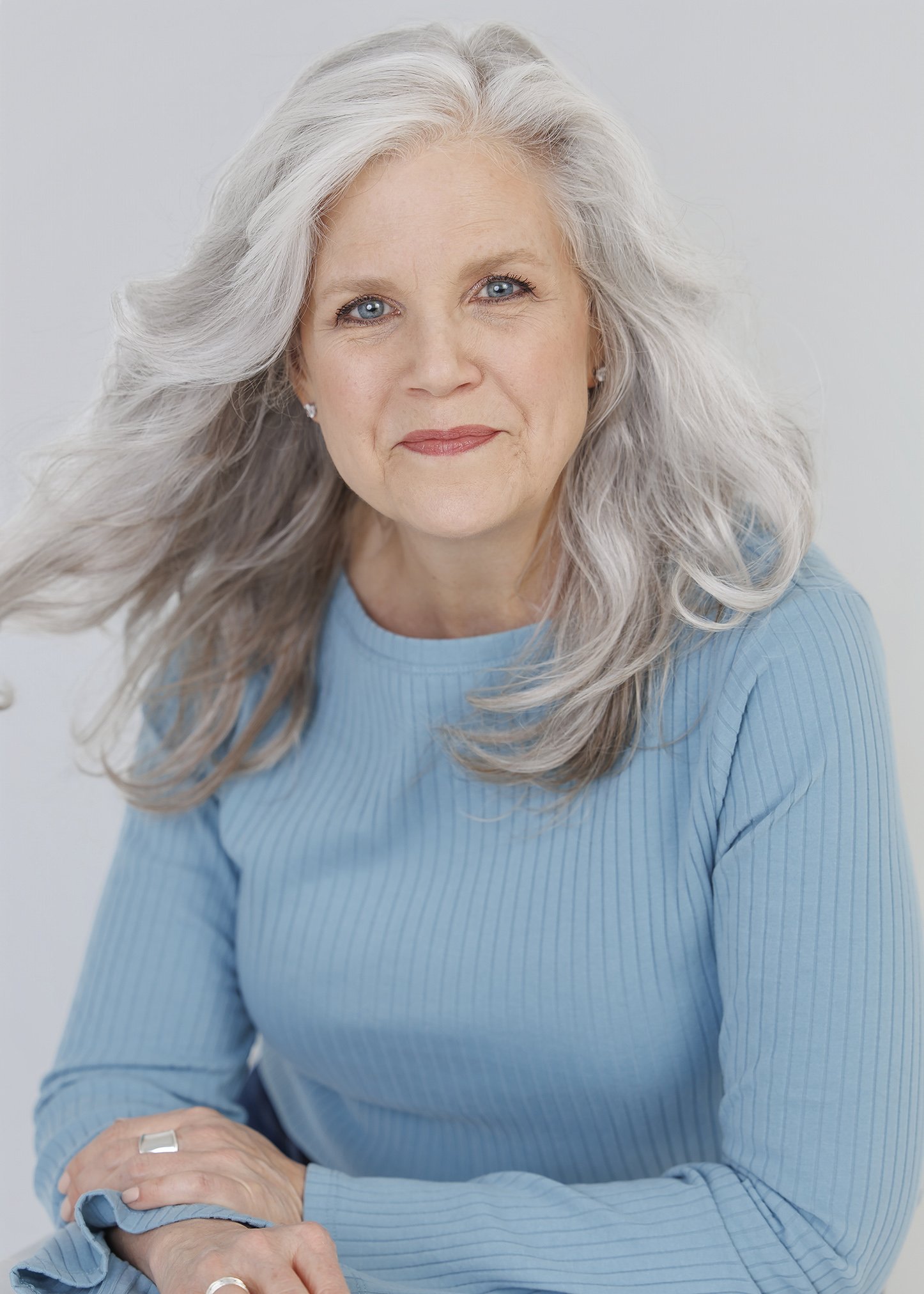 A portrait of an older woman with long, wavy gray hair, blue eyes, and a gentle smile, wearing a light blue ribbed long-sleeve shirt, with her hands resting on a surface, against a plain light gray background.