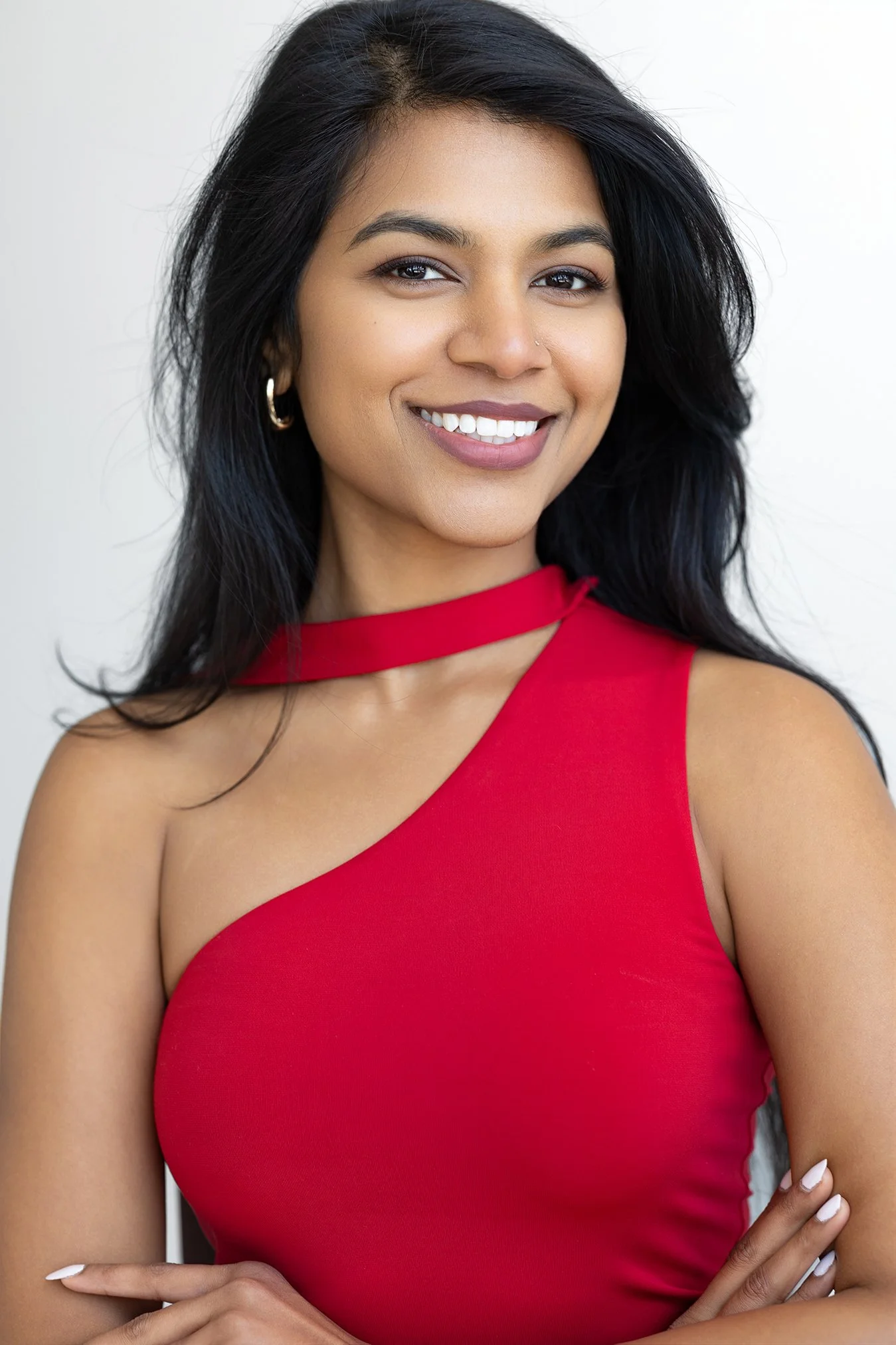 A smiling woman with long black hair, wearing a sleeveless red dress and gold hoop earrings, stands against a white background.