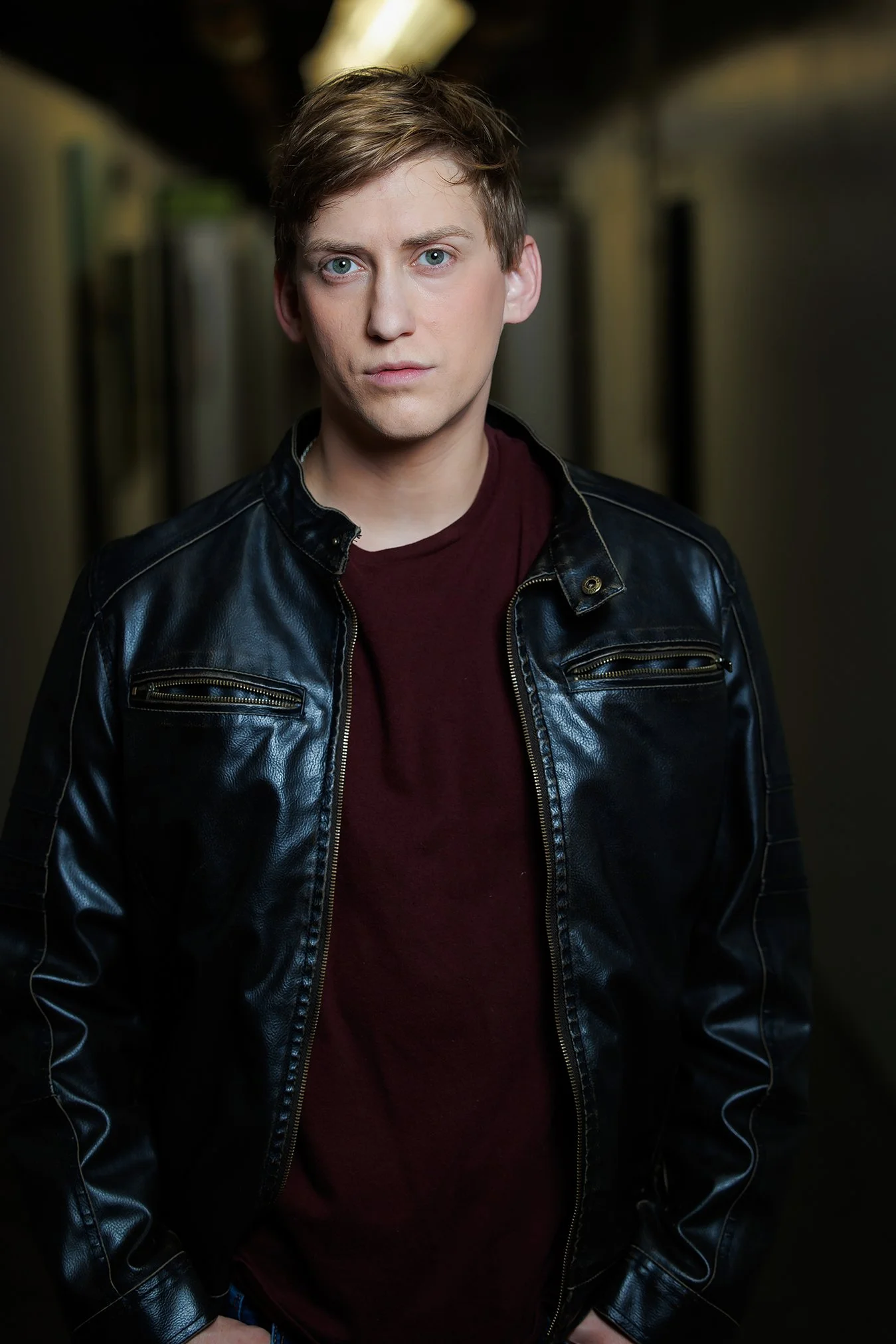 A young man with blonde hair and blue eyes wearing a black leather jacket and a maroon t-shirt, standing in a dimly lit hallway.