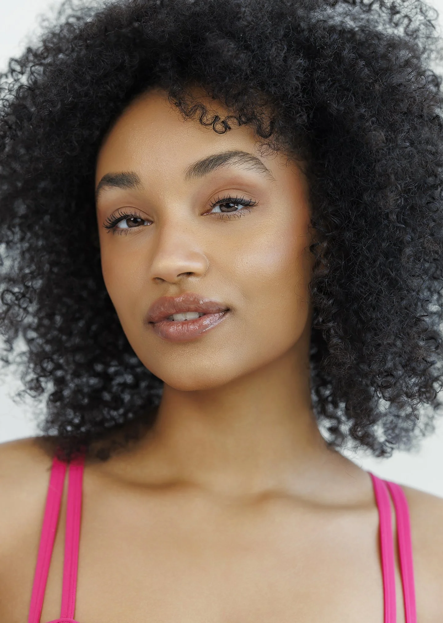 A young woman with natural curly hair wearing a pink spaghetti strap top in a close-up portrait.
