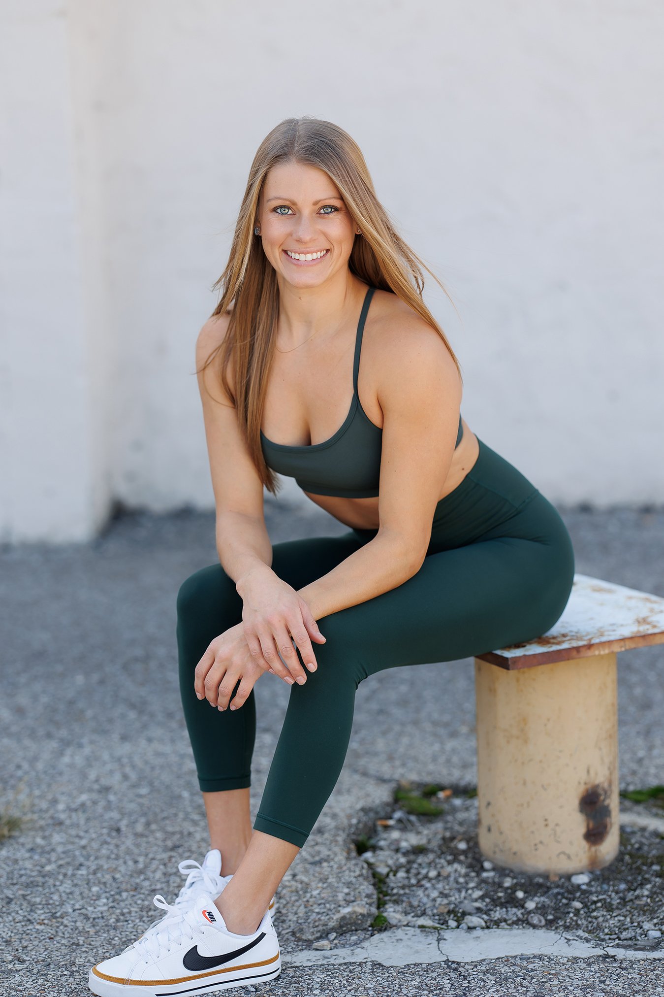 A woman with long hair, wearing a black sports bra and matching leggings, sitting on a bench outdoors. She is smiling and looking at the camera, with a white wall in the background and wearing white Nike sneakers.