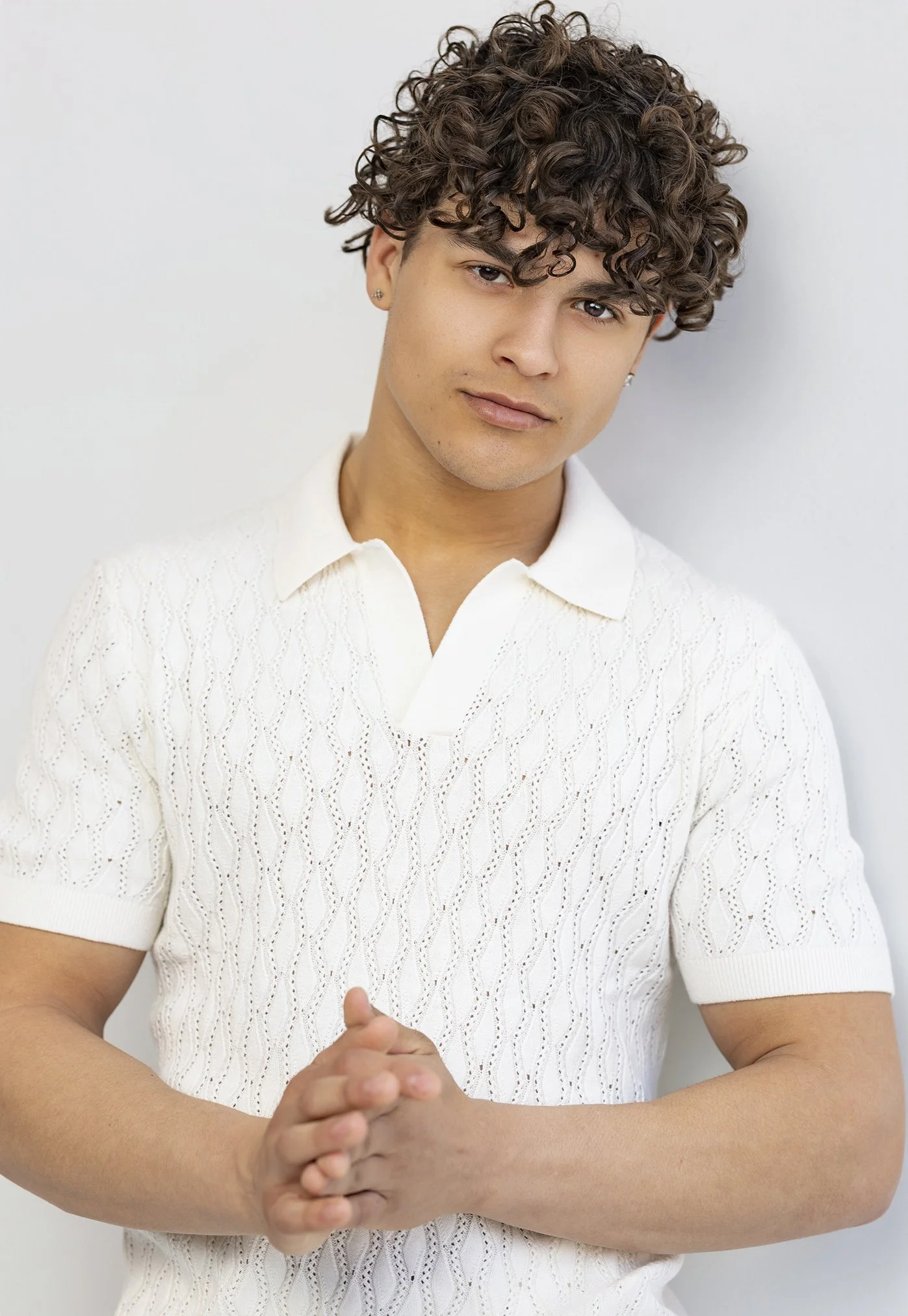 A young man with curly brown hair and earrings wearing a white textured short-sleeve polo shirt standing against a plain white background with hands clasped.