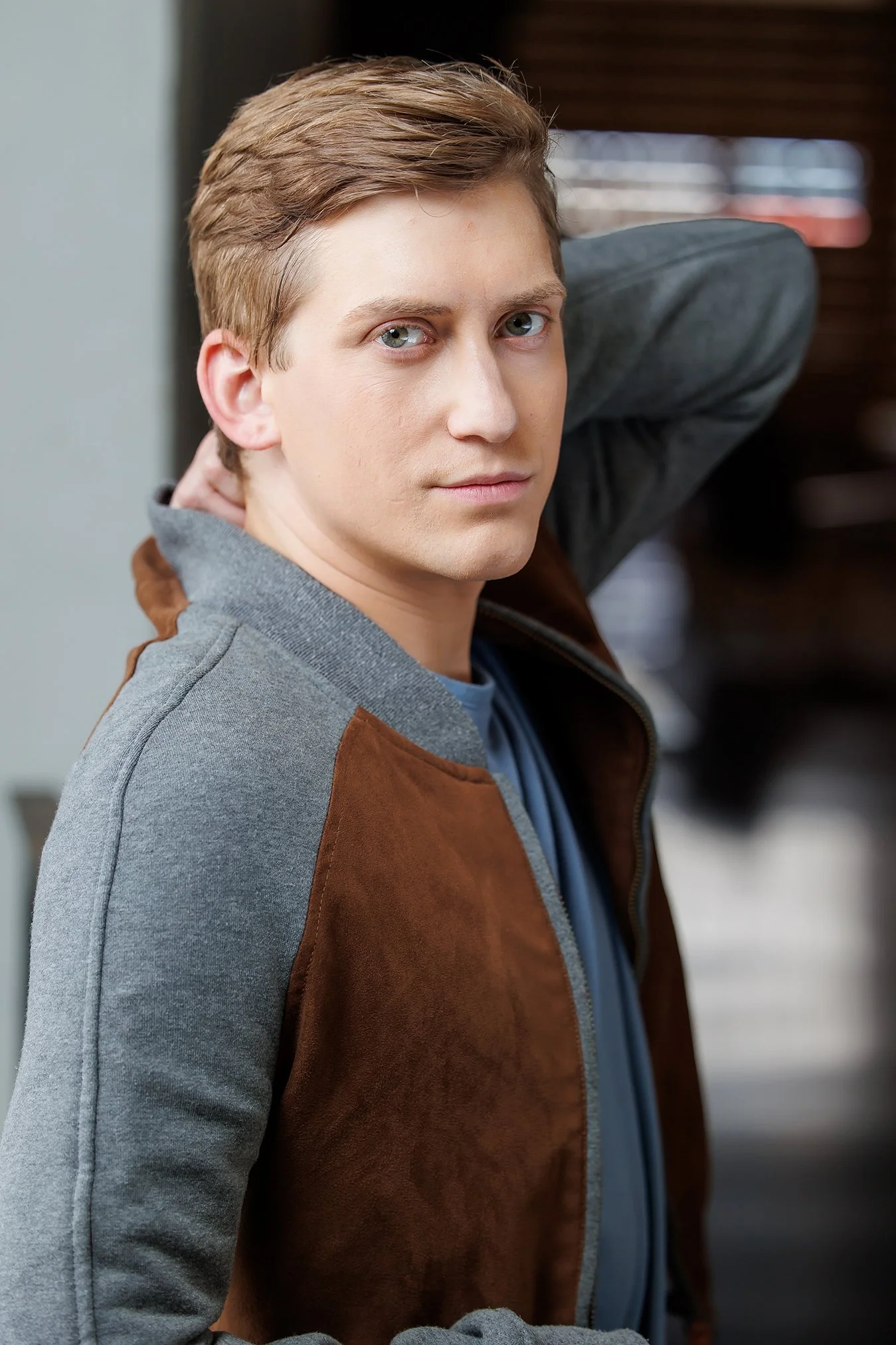 A young man with blond hair and blue eyes, wearing a gray jacket with brown suede sleeves, looking into the camera with a neutral expression, standing indoors.