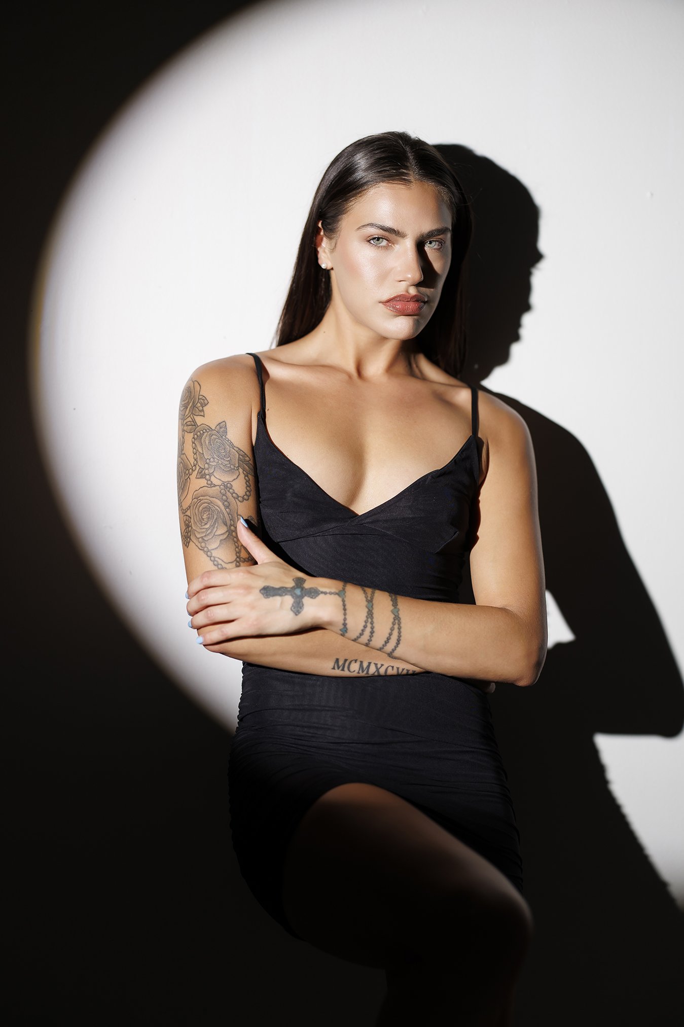 A woman with dark hair and tattoos on her left arm and waist, wearing a black spaghetti strap dress, standing against a white background with a shadow cast on the wall.