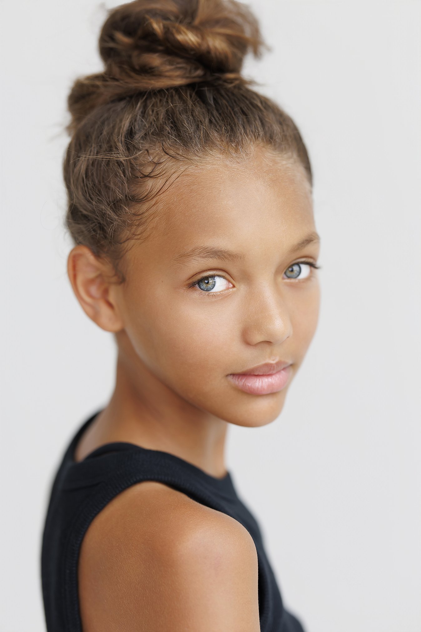 Portrait of a young girl with light brown hair in a top knot, wearing a black sleeveless shirt, looking at the camera with a neutral expression.