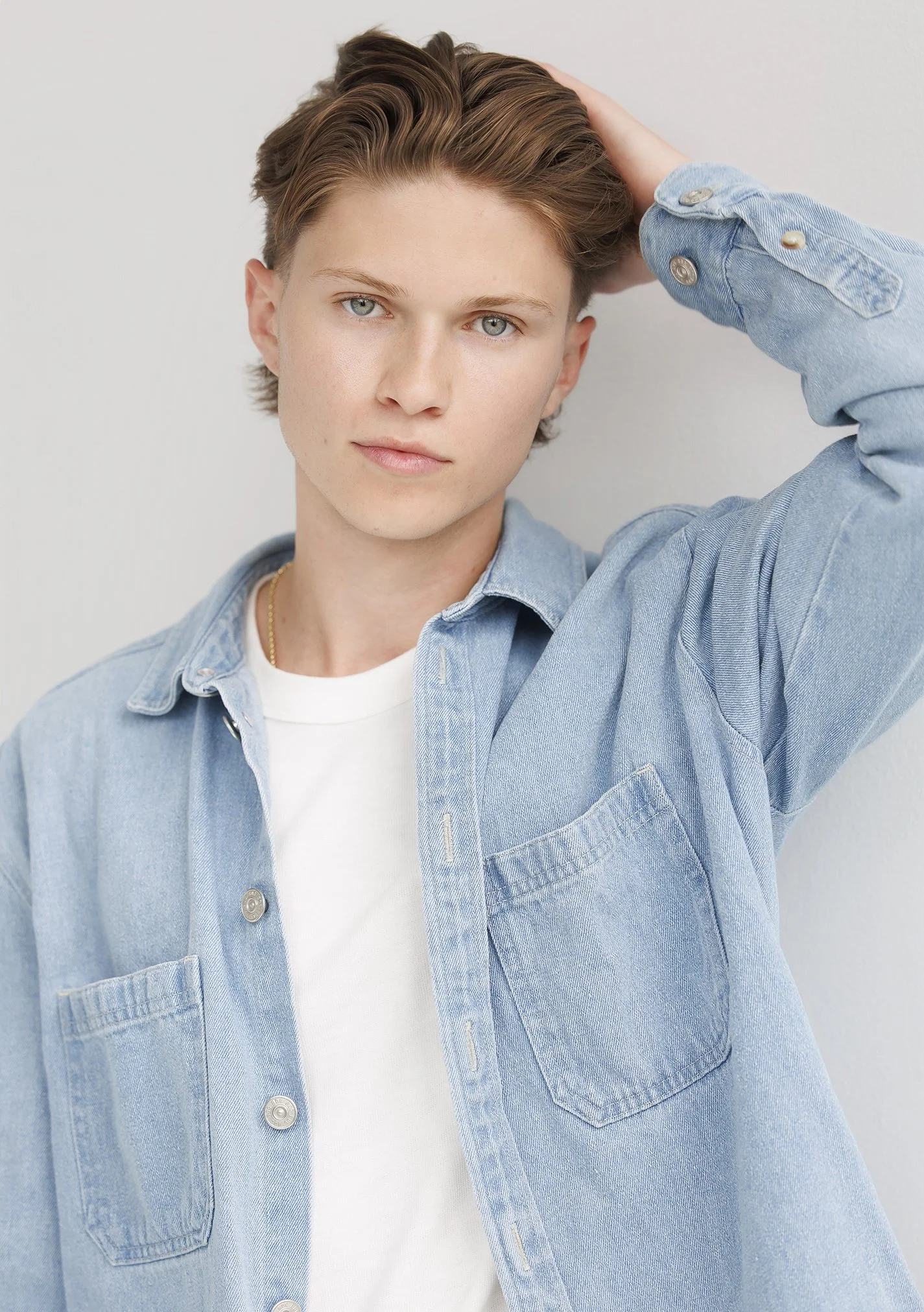 A young man with short, light brown hair and blue eyes, wearing a light denim shirt over a white t-shirt, standing against a plain light background, with his right hand touching the back of his head.