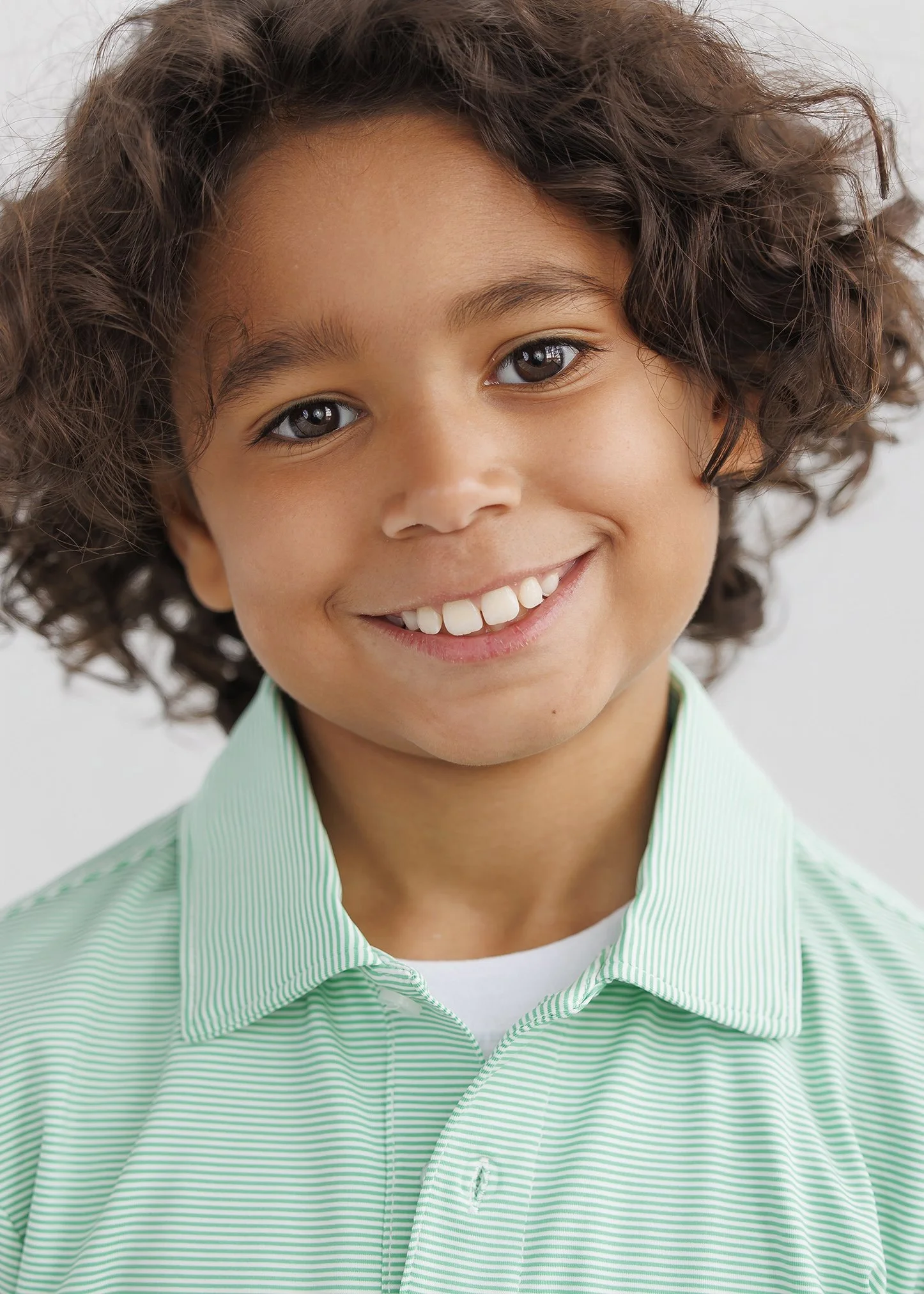 Close-up of a young boy with curly brown hair, smiling, wearing a light green striped button-up shirt.
