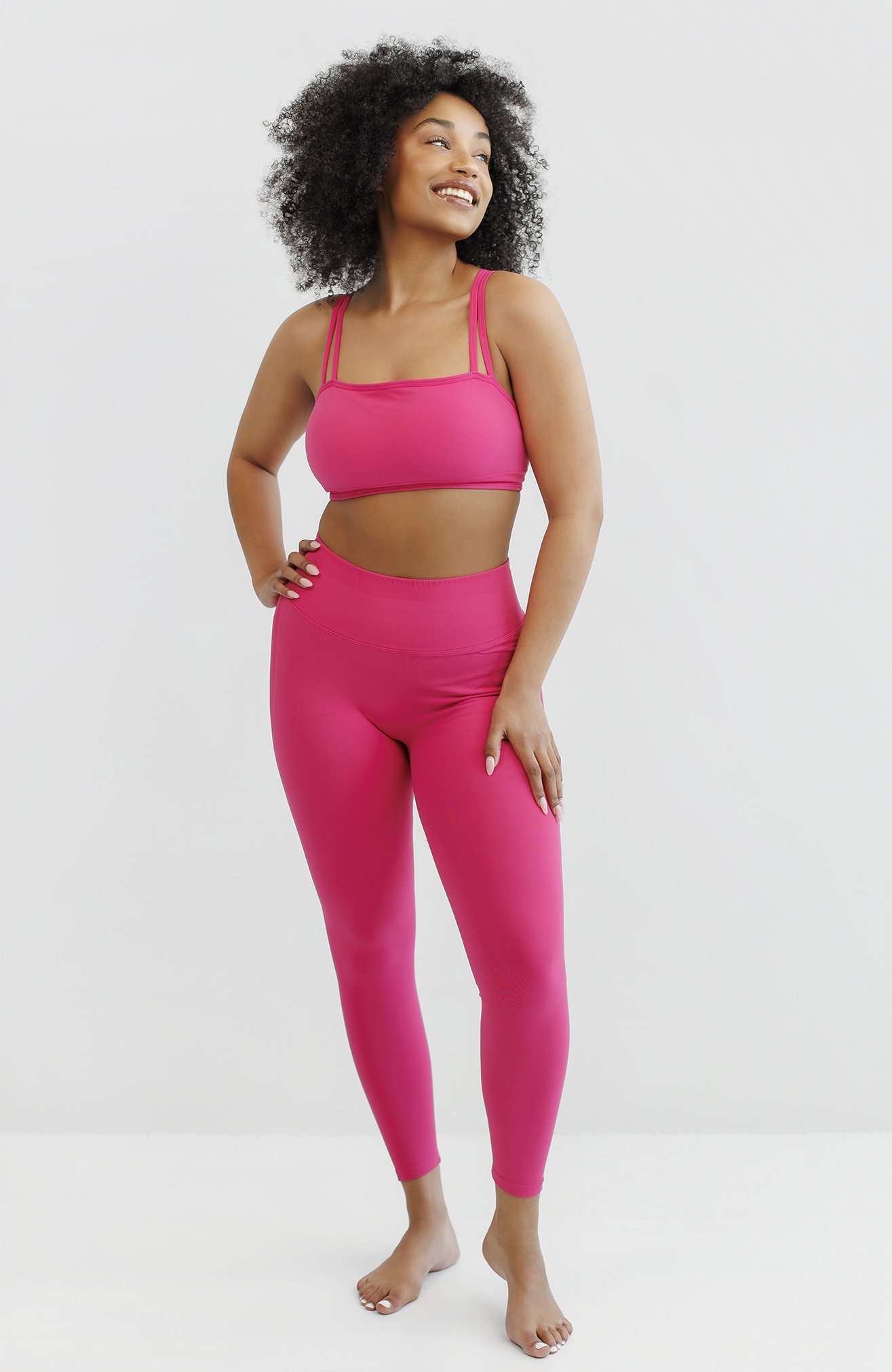 A woman with curly hair in bright pink workout clothes, smiling and standing against a plain white background.