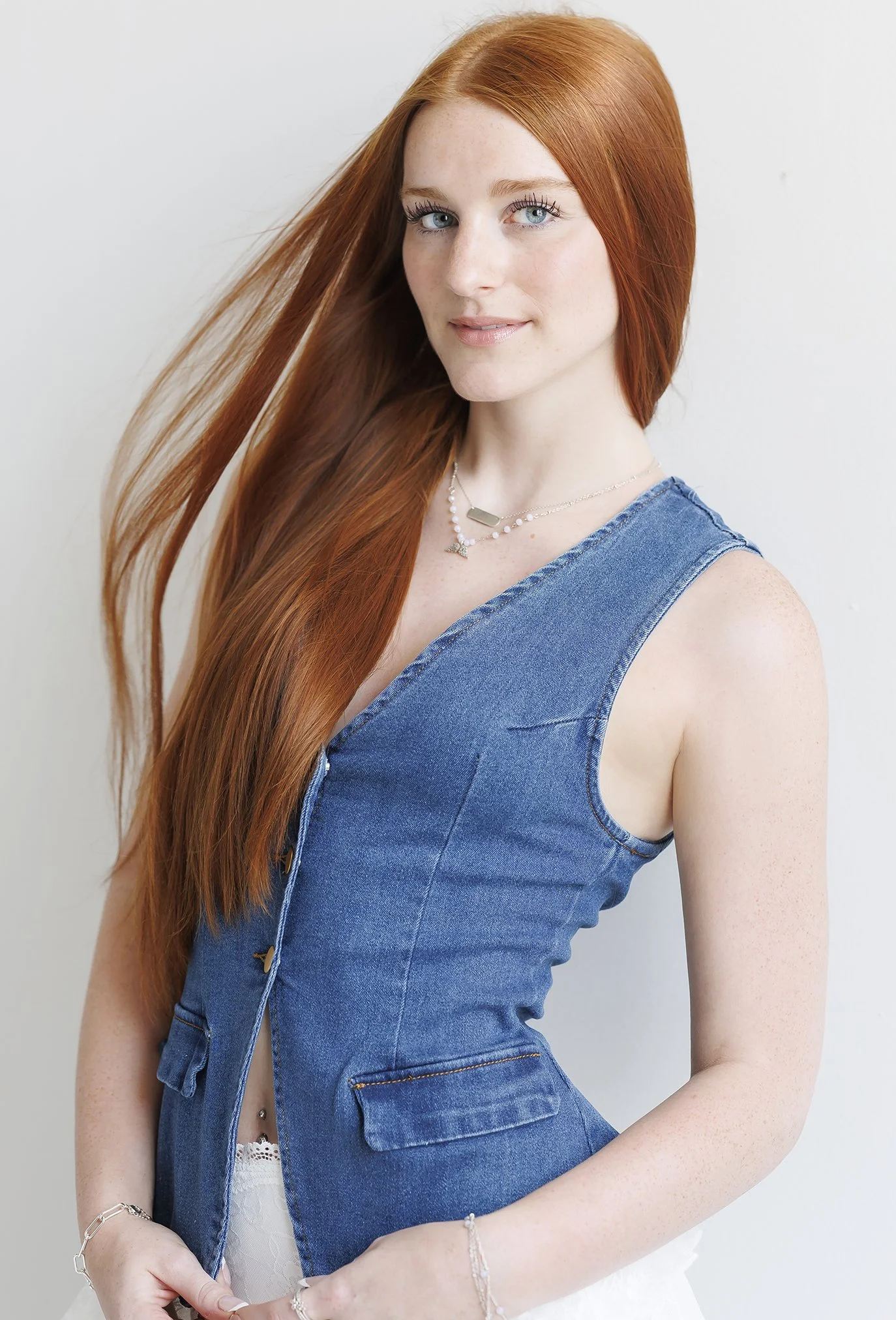 A young woman with long red hair, blue eyes, and fair skin, wearing a sleeveless denim vest and jewelry, standing against a plain white background.