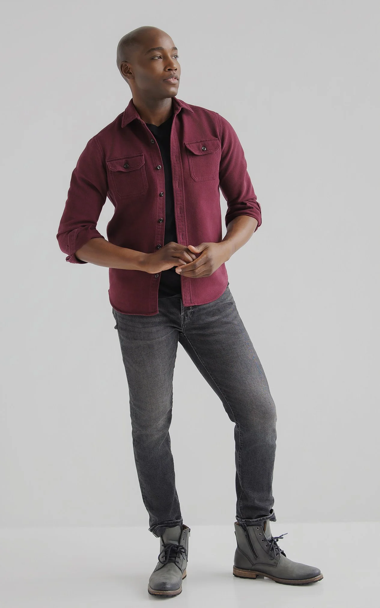A person with a shaved head wearing a burgundy button-up shirt, black shirt underneath, gray jeans, and gray combat boots, standing against a plain light gray background, looking slightly to the side.