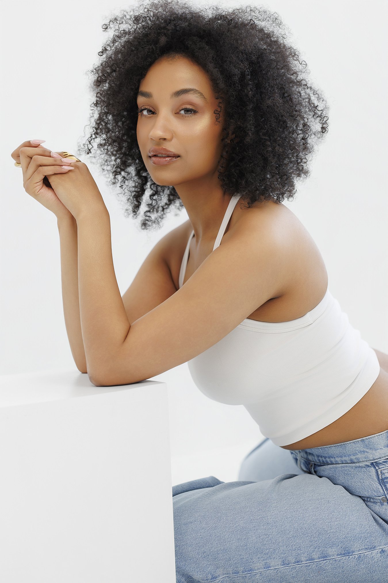 A young woman with curly hair, wearing a white tank top and blue jeans, sitting against a white surface and looking at the camera.