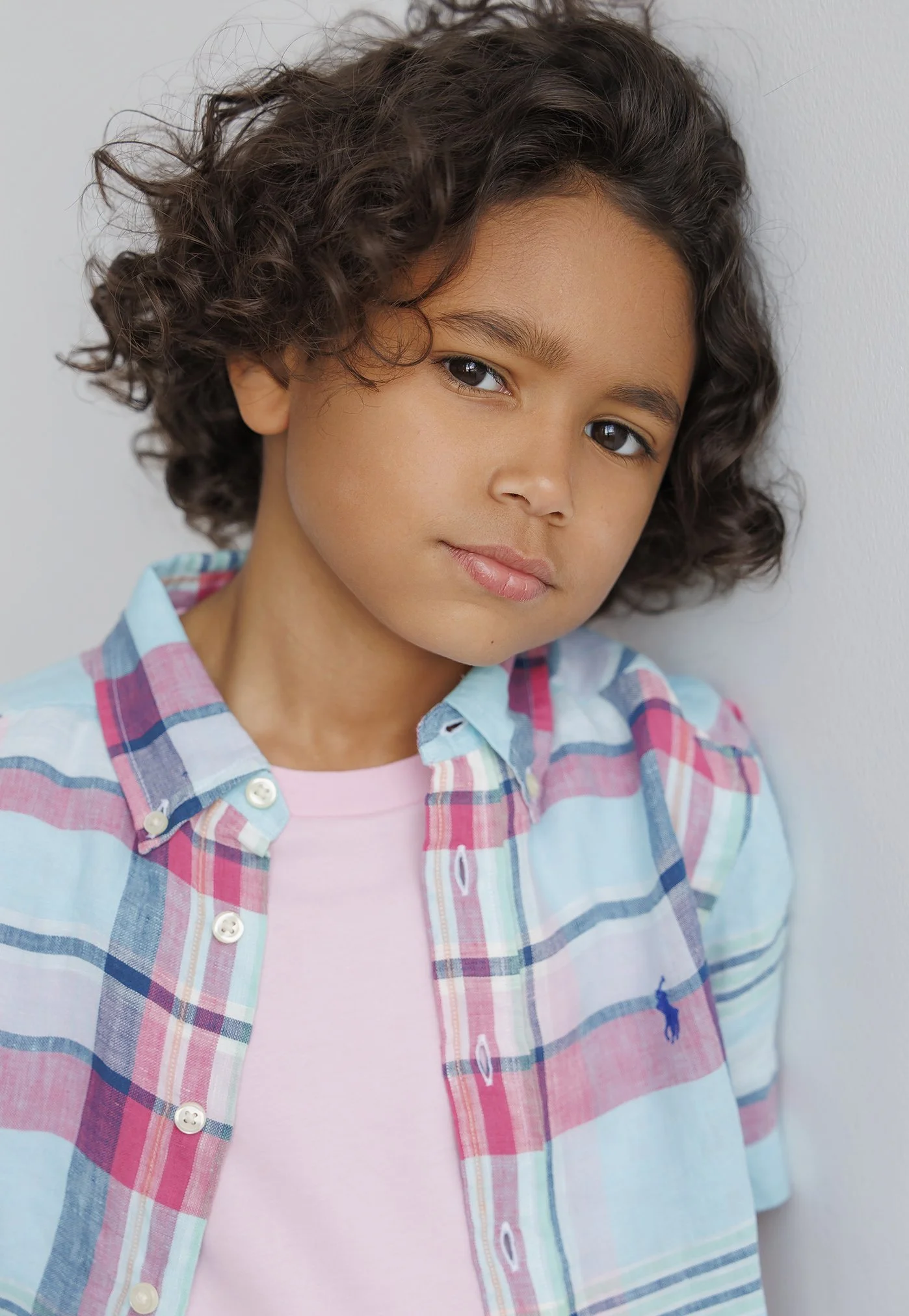 Young boy with curly hair leaning against a wall, wearing a pastel plaid shirt over a pink t-shirt.