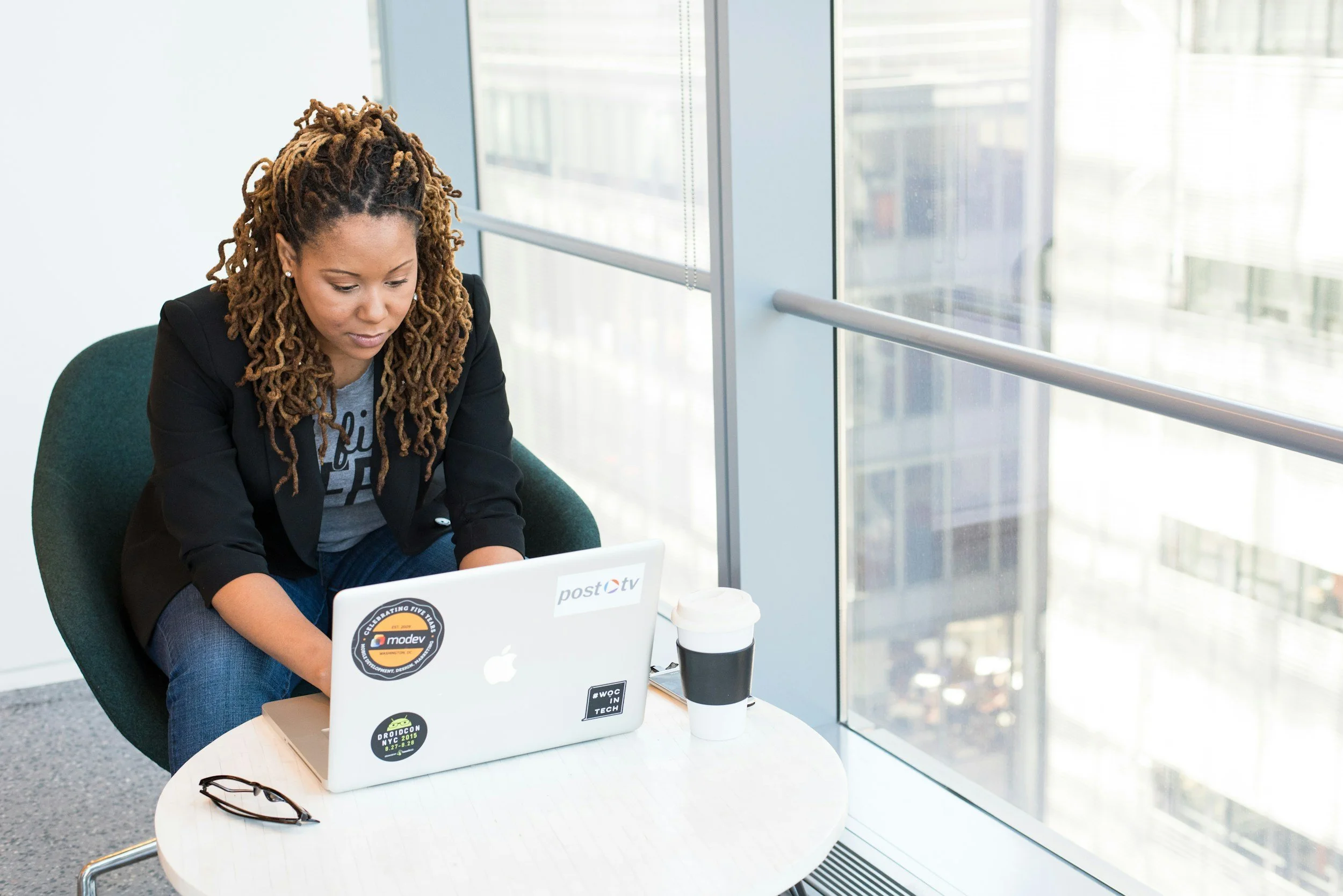 A woman with dreadlocks, wearing a black blazer and gray T-shirt, sitting at a white table with a silver laptop, in front of a large window in a modern office building.