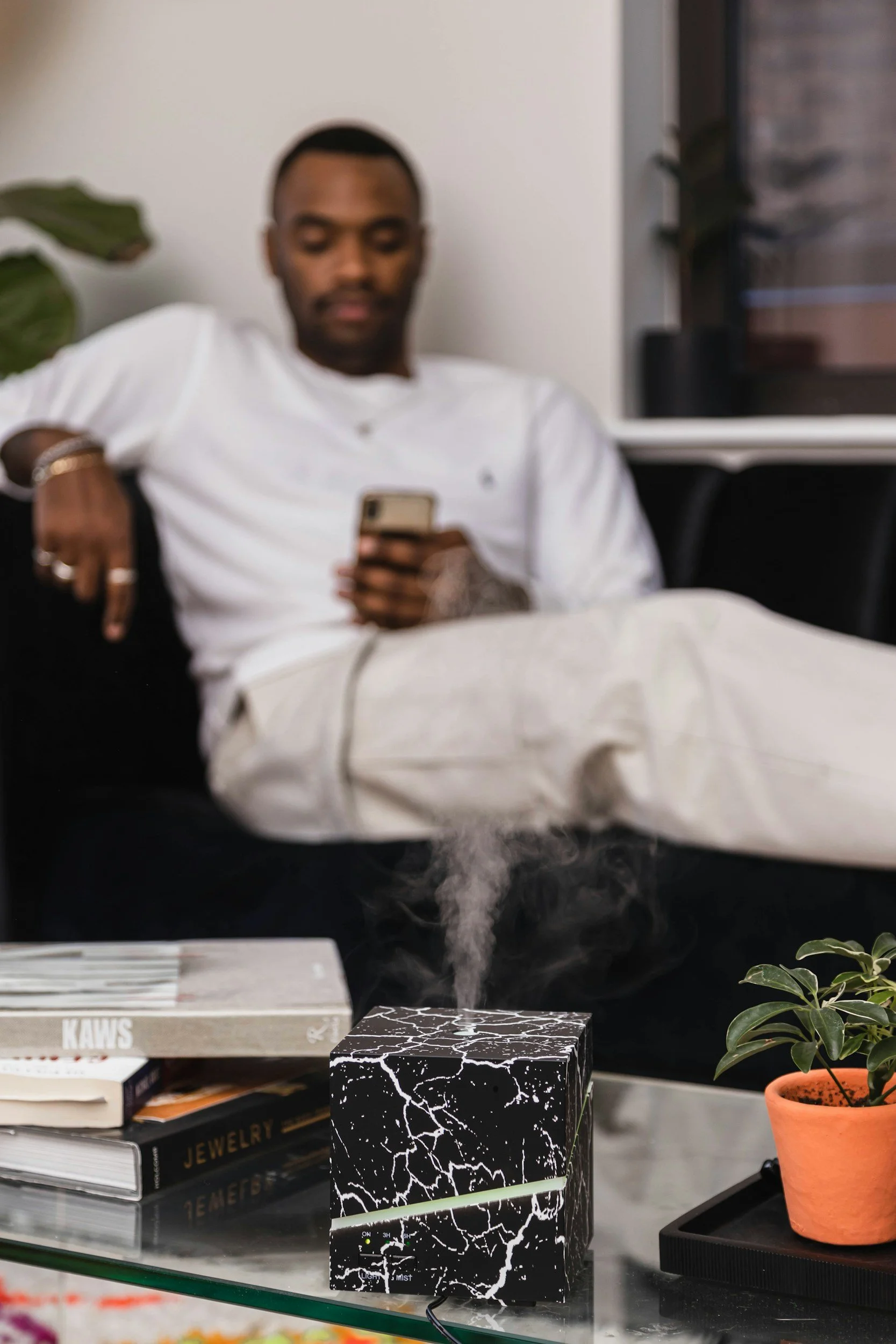 A man in white clothing relaxing on a black sofa, looking at his phone, with a table in front of him holding books and a black and white patterned diffuser