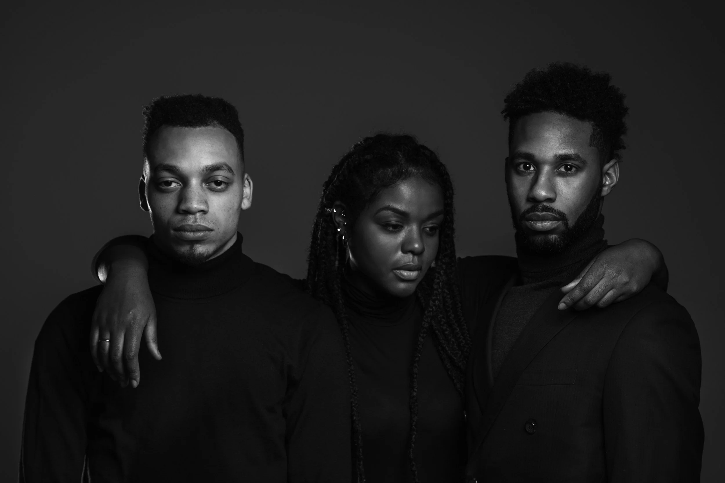 Black and white portrait of three young adults, two men and one woman, with the woman and the man on the right having their arms around each other's shoulders. All three are wearing dark turtleneck sweaters and look directly at the camera with serious expressions.